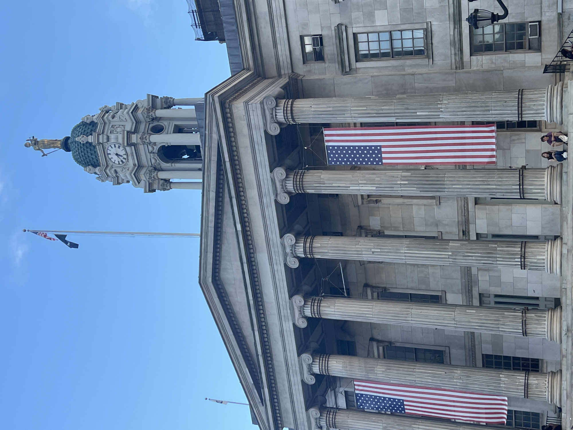 An American flag hangs from the facade of a large stone building with classical architecture, featuring large columns and a clock tower topped with a statue of Lady Justice. Two people are seated on the steps in front of the building. The building appears to be a government or courthouse building. The sky is clear and blue. The location is the Kings County Supreme Court building in Brooklyn, New York City, USA.