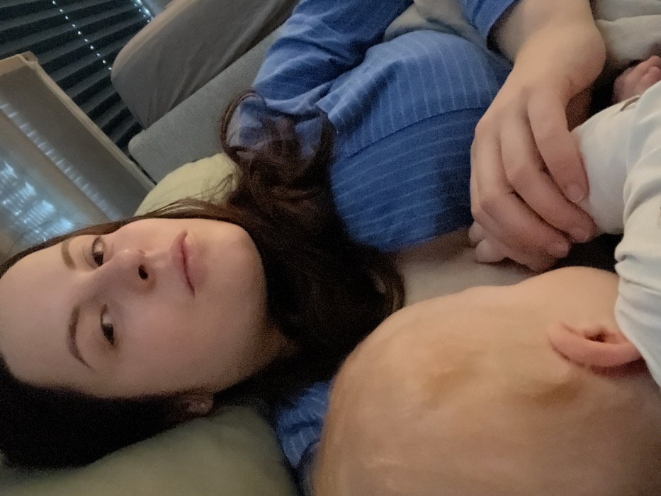 An image depicts an adult female lying down in an indoor setting, looking directly towards the viewer. Her dark hair is spread around her head and shoulders. She wears a blue, long-sleeved garment with faint horizontal stripes. An infant is positioned against her chest, partially visible on the left side of the frame, appearing to be engaged in breastfeeding. The infant's head is largely out of focus, showing light-toned skin and part of an ear. One of the adult female's hands is visible, resting near the infant. She is supported by a light green pillow and rests on a grey fabric surface, likely a sofa or bed. In the background, above her head, elements resembling horizontal window blinds and a subtle pattern (possibly string lights or textured surface) are visible. The lighting is subdued. The specific city of Obermoschel, Germany, cannot be determined from the visual information presented in the image.