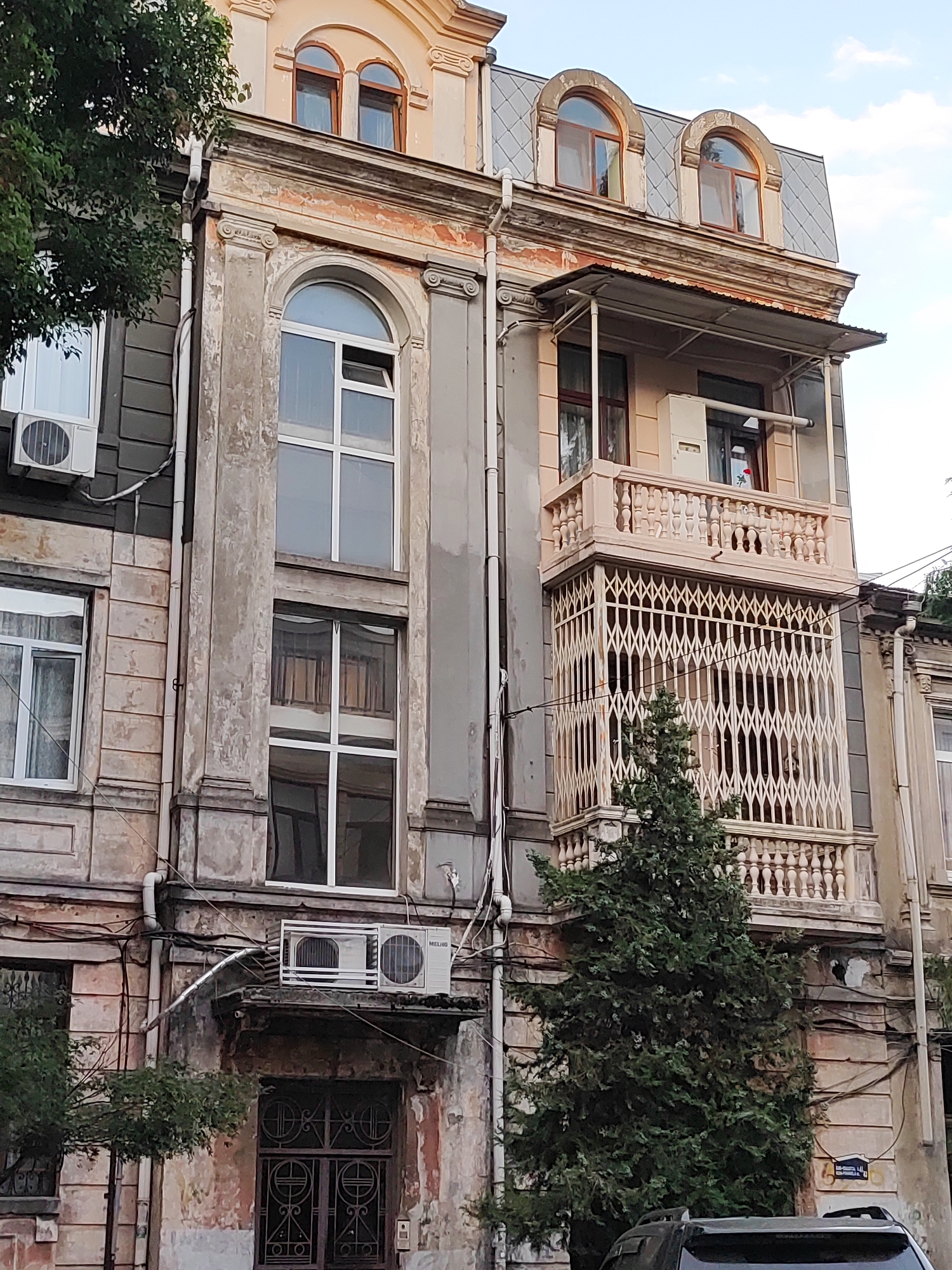 The image shows a historic building in Batumi, Georgia. The building has a weathered facade with multiple windows, some arched and some rectangular. A balcony with ornate railings extends from the upper floor, and a decorative metal grille covers the lower portion of the balcony. An air conditioning unit is visible on the exterior wall. A large evergreen tree is positioned in front of the building. The sky above is partly cloudy. The overall impression is one of aged architecture in an urban setting.