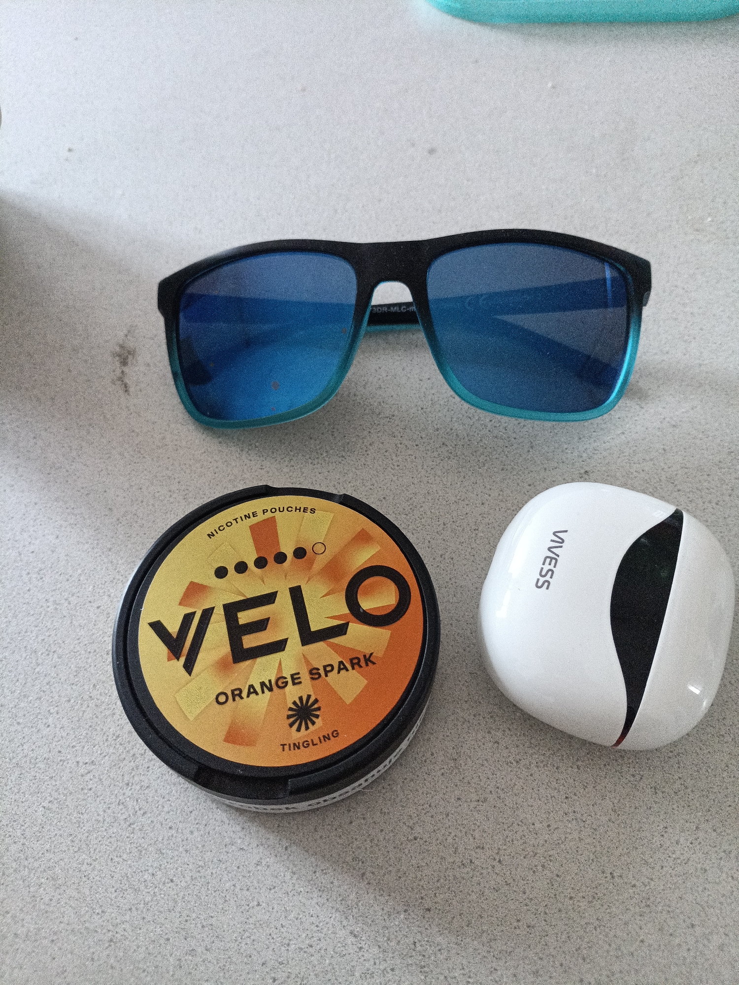 The image shows a top-down view of a light gray, speckled countertop. On the countertop are three items: a pair of black-framed sunglasses with bright blue lenses, a round, orange tin of "VELO Orange Spark" nicotine pouches, and a white, pebble-shaped electronic device with a black accent. The sunglasses are positioned at the top of the frame, angled slightly. The Velo tin is to the left, with its lid prominently displaying the brand name and flavor. The electronic device is to the right of the tin. The lighting suggests an indoor environment, likely a home or office. There is no information in the image that can indicate the location in the city of Dalovy, Czechia.