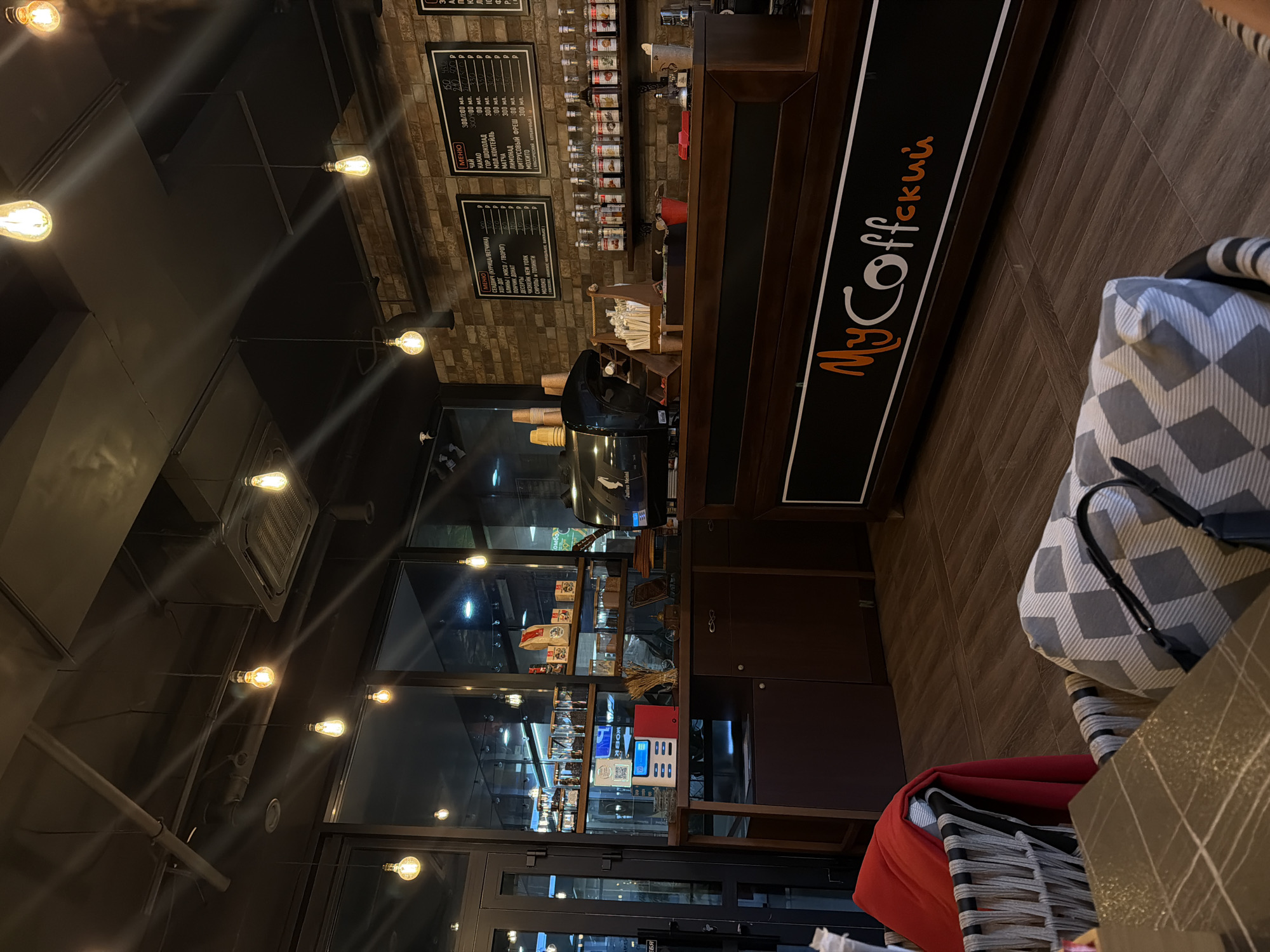Here is a detailed description of the image:
**Scene:** The image shows the interior of a coffee shop called "MyCoffee". It is likely located in the city of Sochi, Russia, given the Cyrillic alphabet used in the coffee shop's name and menu.
**Environment:** The coffee shop has a modern and slightly industrial aesthetic with exposed brickwork and hanging Edison-style light bulbs. The interior is dimly lit, creating a cozy atmosphere.
**Objects:**
* A counter runs along the right side of the image with the "MyCoffee" logo prominently displayed.
* Behind the counter are shelves stocked with coffee beans, syrups, and other coffee-related items.
* A glass display case is visible, possibly containing pastries or other food items.
* A menu board is mounted on the wall behind the counter with prices in rubles
* A cash register
* A gray and white patterned armchair is visible in the foreground, with a black handbag hanging off of it.
* A small gray table
* A red pillow on a chair in the lower right.
**People:** There is a person at the counter, possibly a customer.
**Interactions:** A transaction appears to be occurring at the counter, although the specifics are unclear.
**Overall Impression:** The image captures a moment inside a local coffee shop in Sochi. It conveys a sense of warmth and invitation.