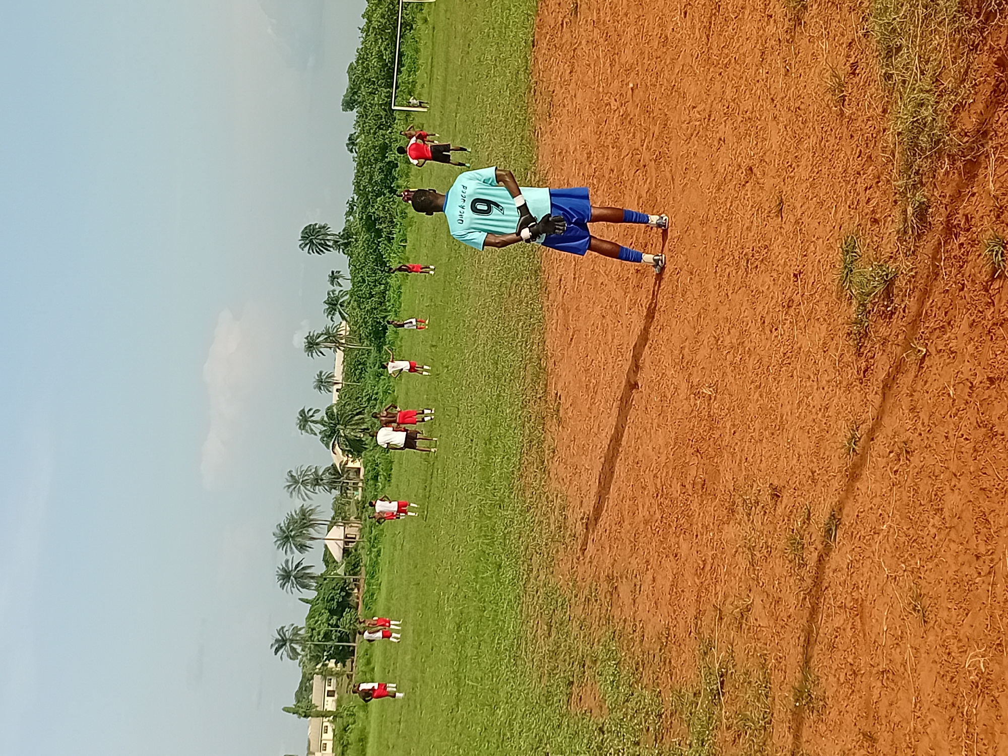 A soccer game is in progress on a grassy field in Ogbogbo, Nigeria. One player, wearing a light blue jersey with the number 9 on the back, is standing on a dirt area adjacent to the field. Other players in red and white jerseys are visible on the field. Palm trees are visible in the background. Buildings are visible in the distance. The sky is light blue with some clouds.