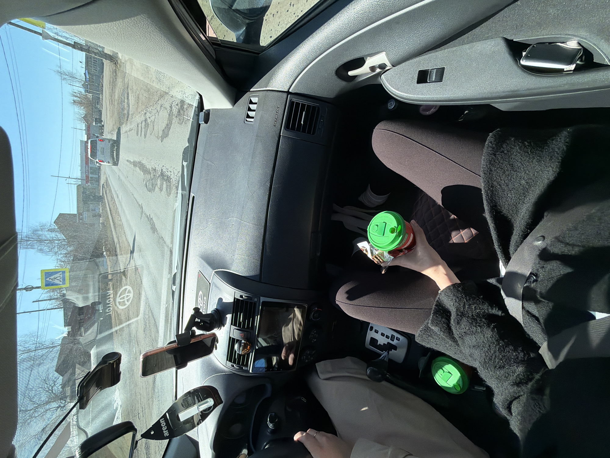 The scene is the interior of a car, seemingly a Toyota, traveling along a street in what appears to be Chaikovsky, Russia, as suggested by the street signs and architecture visible through the windshield. The road ahead shows signs of wear. The driver is behind the steering wheel, and a passenger is in the front seat. The passenger is holding a green-lidded drink container and appears to be eating something. Another similar drink container is located near the center console. Various objects are mounted on the windshield, including a phone holder and what appears to be an air freshener. A passenger's seat is black and quilted, with someone sitting in it wearing black clothing and a seatbelt.