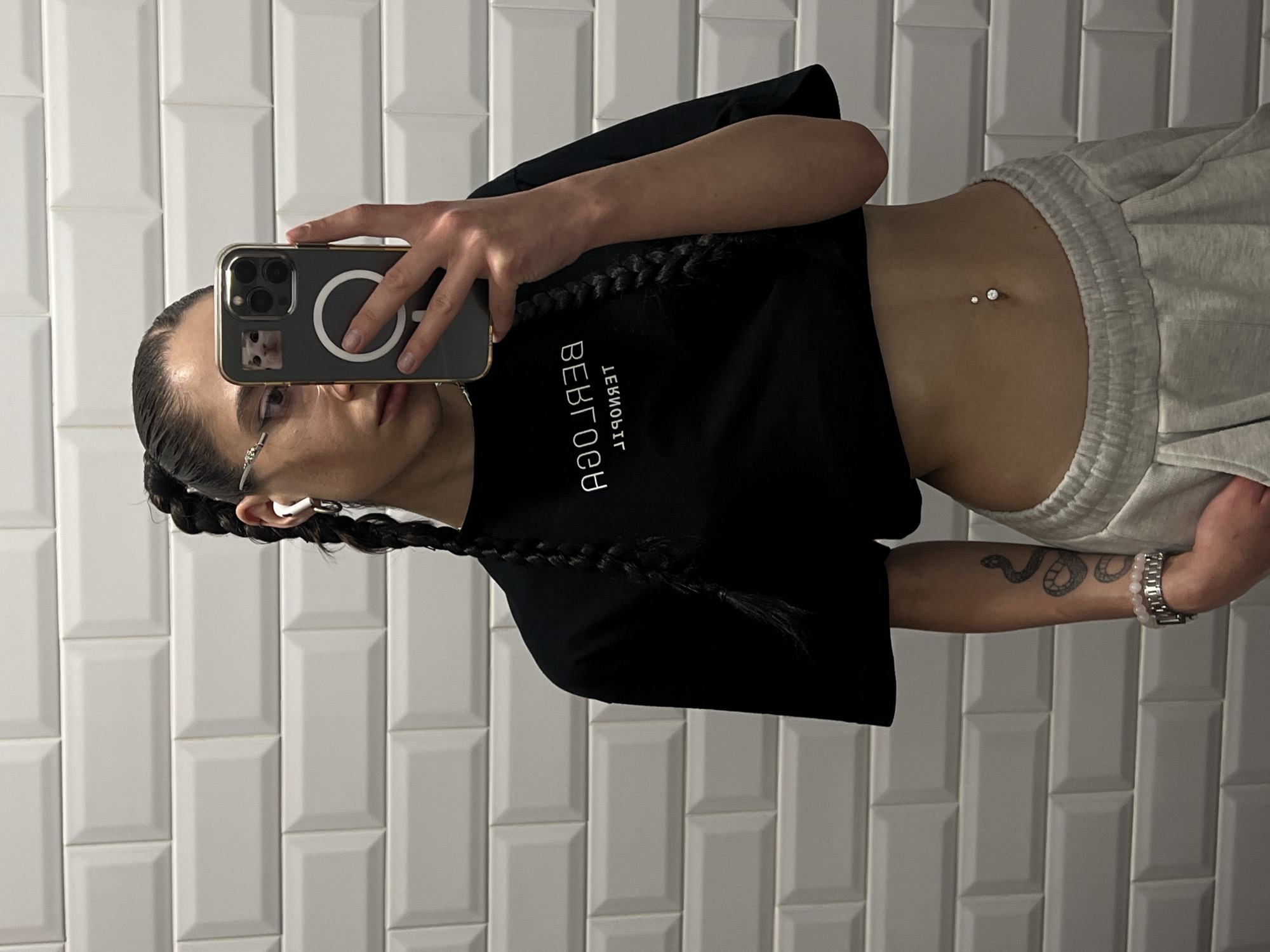 A young woman with dark hair in two braids and wearing glasses takes a mirror selfie. She is dressed in a black crop top and light-colored sweatpants. Her midriff is exposed, revealing a navel piercing with two small gems. A snake tattoo is visible on her right arm, along with a silver watch with a pink beaded bracelet. She is holding a smartphone with a gold case, which is reflecting her face and the phone's screen, showing a cat image. The background consists of white subway tiles. The image appears to be taken indoors, possibly in a bathroom, given the tile work. There is no indication of the time of day or weather. The overall mood is casual. Text visible on the woman's top is "BEBTOOB" and "ЛЕЯИОЫЯ".
