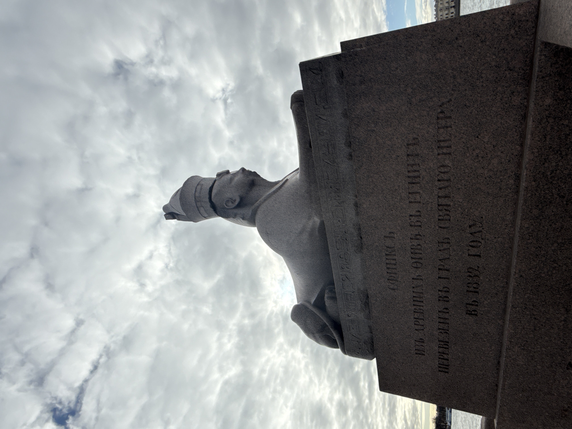 The image shows a sphinx statue in Saint Petersburg, Russia. The statue is made of granite and has a human head and the body of a lion. The sphinx is located on the University Embankment near the Mining Institute. There is a text inscription on the pedestal. The sky is cloudy. The image is taken from a low angle.