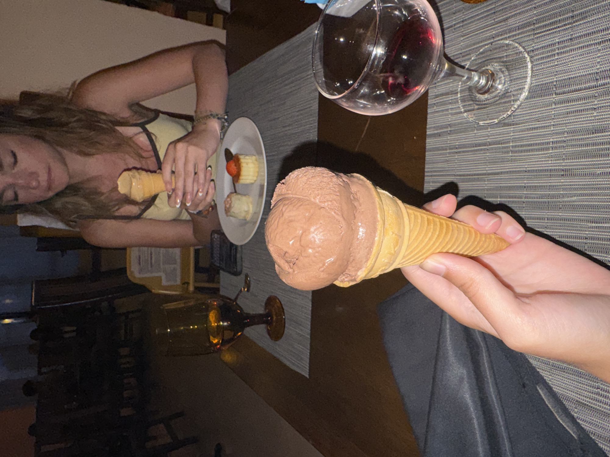 A woman and person hold ice cream and dessert on a table with drinks. The photograph appears to have been taken in Playa del Carmen, Mexico. A woman with long hair and a tank top holds a small ice cream cone. She is seated at a table near a white plate with a small cake-like dessert topped with fruit. A second person holds a larger chocolate ice cream cone. A partially-filled wine glass and a glass of amber liquid are on the table as well. The table has a grey textured surface. The overall lighting is dim.