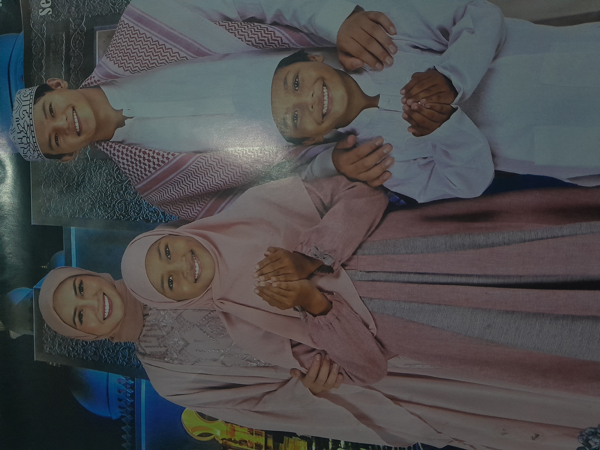 The image depicts a family of four posing for a photograph, likely for a religious or cultural occasion, given their attire. In the foreground, a young boy in a white tunic and cap holds his hands together in prayer. To his left, a young girl in a pink hijab and dress also has her hands clasped in prayer. Behind them, a woman wearing a light pink hijab and dress stands with her arm around the girl, and a man in a white tunic and a red and white checkered headdress stands behind the boy, with his arms around the children. The background features a stylized depiction of Islamic architecture, possibly a mosque, with arches and domes illuminated in blue. The overall scene suggests a celebratory or devotional atmosphere. The setting appears to be indoors, possibly a studio or a decorated space designed for photography. While the image lacks specific landmarks to pinpoint its exact location within Depok, Indonesia, the cultural context of the people and their clothing suggests a connection to the Indonesian Islamic community.