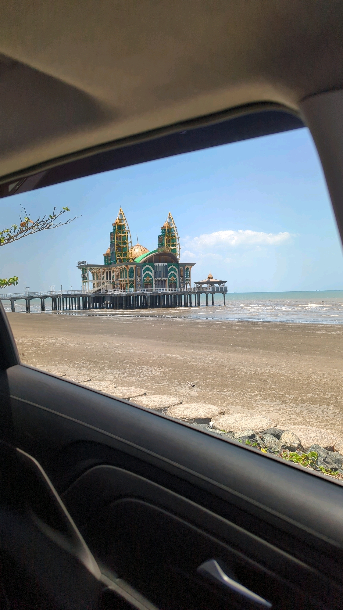 An eye-level, medium outdoor shot captures the Masjid Raya Baiturrahman, also known as the Floating Mosque, located in the city of Beringin, Indonesia. The mosque is a prominent architectural structure situated over the ocean, supported by numerous pillars. Its design features a large central dome and two towering minarets, all adorned in vibrant green and gold colors. A long walkway extends from the shore to the mosque. The foreground of the image is dominated by the interior of a car, with the car's dashboard and door panel visible. The car's windshield frames the view of the mosque, the ocean, and a sandy beach. The sky is a clear, bright blue with a few scattered clouds. The water is calm, with gentle waves lapping the shore. The beach is composed of light brown sand, interspersed with some rocks and small green plants near the waterline. The overall scene depicts a serene coastal landscape with a striking religious landmark.