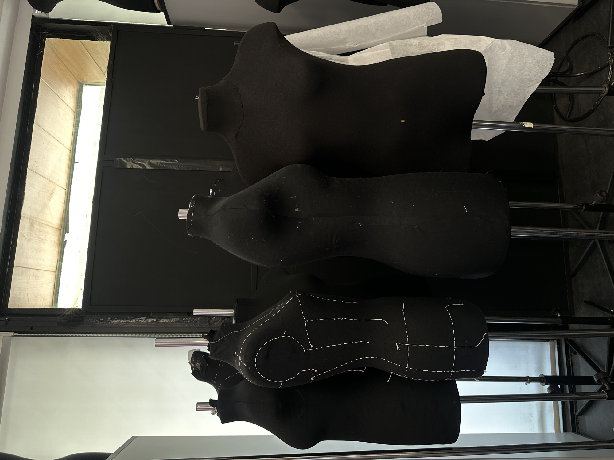 The image shows a collection of black dressmaker's dummies, stacked vertically on metal stands. The dummies vary in size and shape, with one displaying white basting stitches outlining a pattern. Behind the dummies, a window provides natural light, illuminating a wall with light-colored panels or siding visible through the glass. The background appears to be a studio or workshop environment. The image contains no people or subjects.
I have no information regarding the location of this image being in the city of Moscow, Russia.
