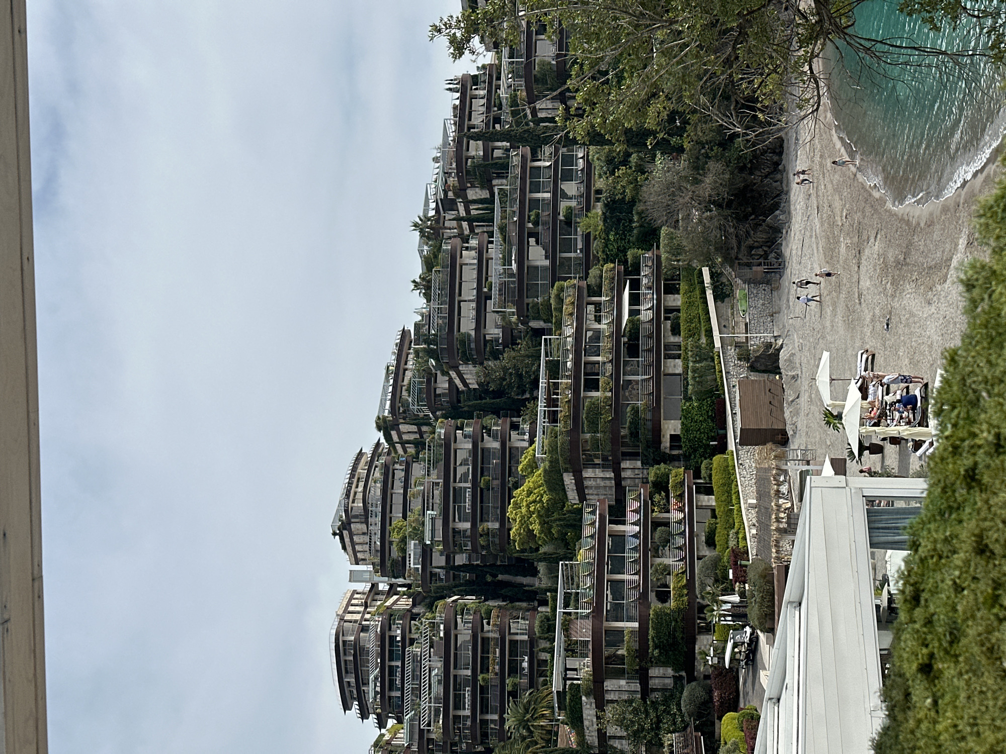 The image depicts a beach scene in Budva, Montenegro, with modern apartment buildings and a sandy beach. The buildings, stacked in tiers on a hillside, feature balconies with greenery and decorative metal railings. The beach is light brown, with a few people walking and some relaxing on sun loungers under umbrellas. Clear turquoise water laps the shore, suggesting a calm sea. The sky is overcast with a flat gray color. Vegetation is visible throughout the scene, with trees interspersed among the buildings and along the beach.
