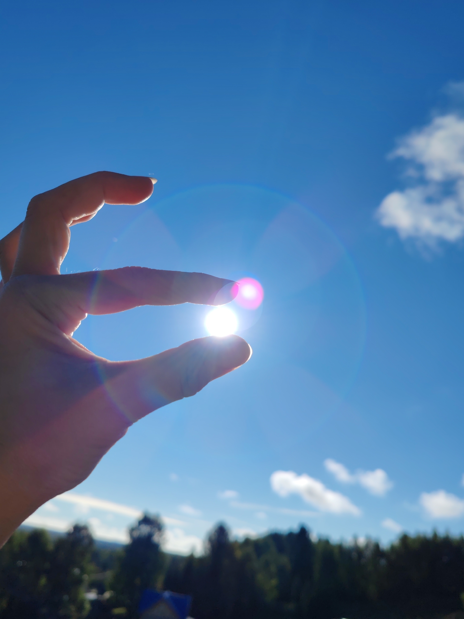 A close-up, low-angle shot captures a person's hand silhouetted against a bright blue sky with scattered white clouds. The sun is visible, creating a lens flare that casts rainbow-colored light across the image. The person's thumb and index finger are held close together, appearing to pinch the sun. In the blurred background, a line of dark green trees is visible, with a hint of a blue roof and some houses at the bottom of the frame. The image was taken in the city of Chukhlem, Russia.