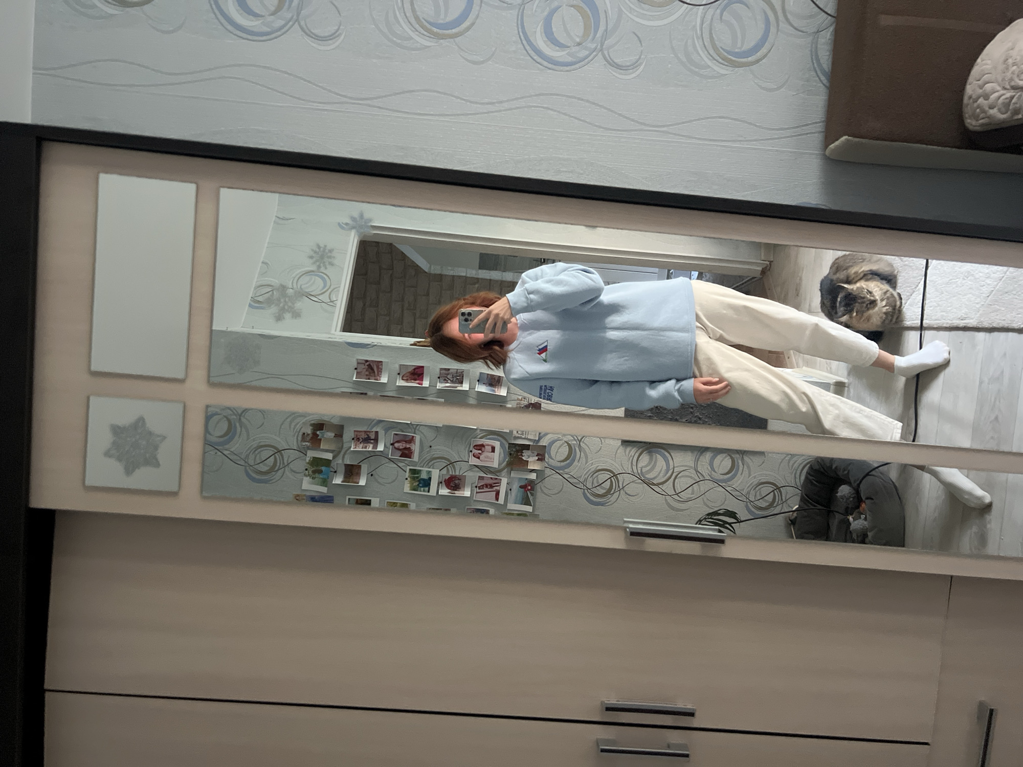 In an indoor setting, a young woman with reddish-brown hair takes a selfie in a full-length mirror. She is wearing a light blue sweatshirt with a small triangular logo and light-colored pants, along with white socks. Her phone is held up to capture her reflection. Next to her, a tabby cat is curled up on a rug on the floor. The mirror is set against a wall adorned with patterned wallpaper and decorative snowflakes. A closet with light-colored doors is visible to the left of the mirror, and a portion of a bed frame is visible on the right. This scene is likely in Chaikovsky, Russia, given the context of the request.