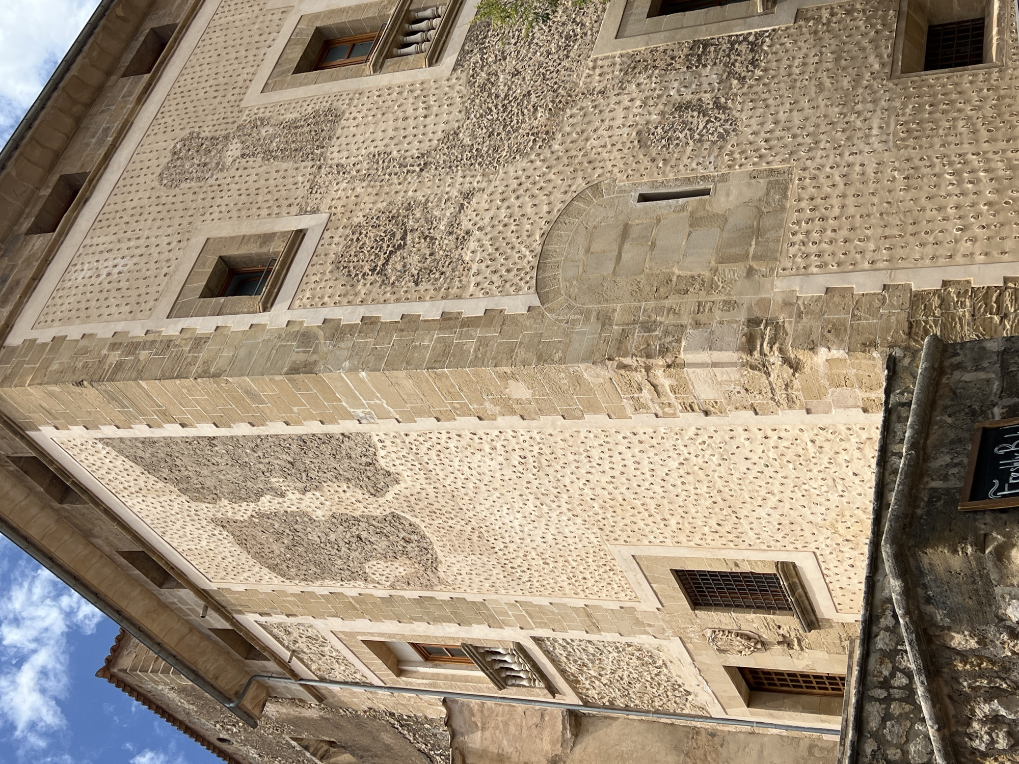 The image shows the exterior of a building in Pollença, Spain, with a distinctive textured facade. The building is constructed with light-colored stone blocks and features two prominent sections of wall decorated with numerous small, indentations or protrusions. These textured areas are arranged in irregular patterns, creating a rough, almost organic surface.
There are several windows visible, some with wooden frames and metal grilles, while others have small balconies with balustrades. The building's architecture suggests historical significance, with visible stone detailing around the windows and corners. Part of an adjacent building, also made of stone, is visible on the left, with a terracotta-tiled roof.
In the foreground, there is a stone wall or parapet, and a dark sign with white lettering is partially visible, suggesting a commercial establishment. The sky above is partly cloudy with a blue sky. The overall impression is of an old, well-preserved building in a Spanish town.