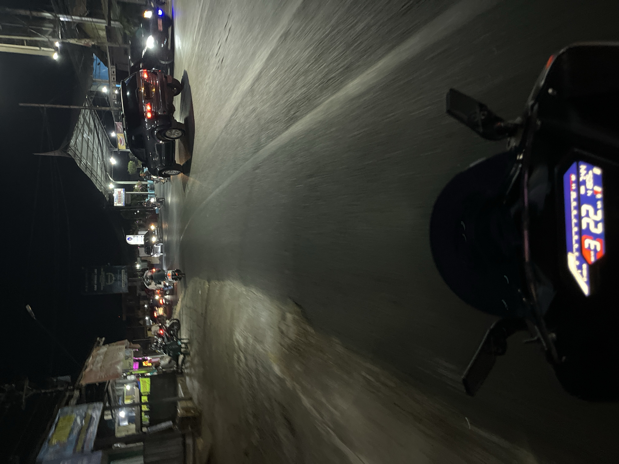A first-person perspective shot from a motorcycle at night in Kalinyamatan, Indonesia. The motorcycle's digital display shows "3 22.". The road is wet and reflects streetlights, indicating recent rain. Traffic includes a black SUV, a white car, and several motorcycles. Buildings with neon signs line the left side of the road. A covered awning structure extends over part of the road in the background. The atmosphere is dark and lit by artificial lights.