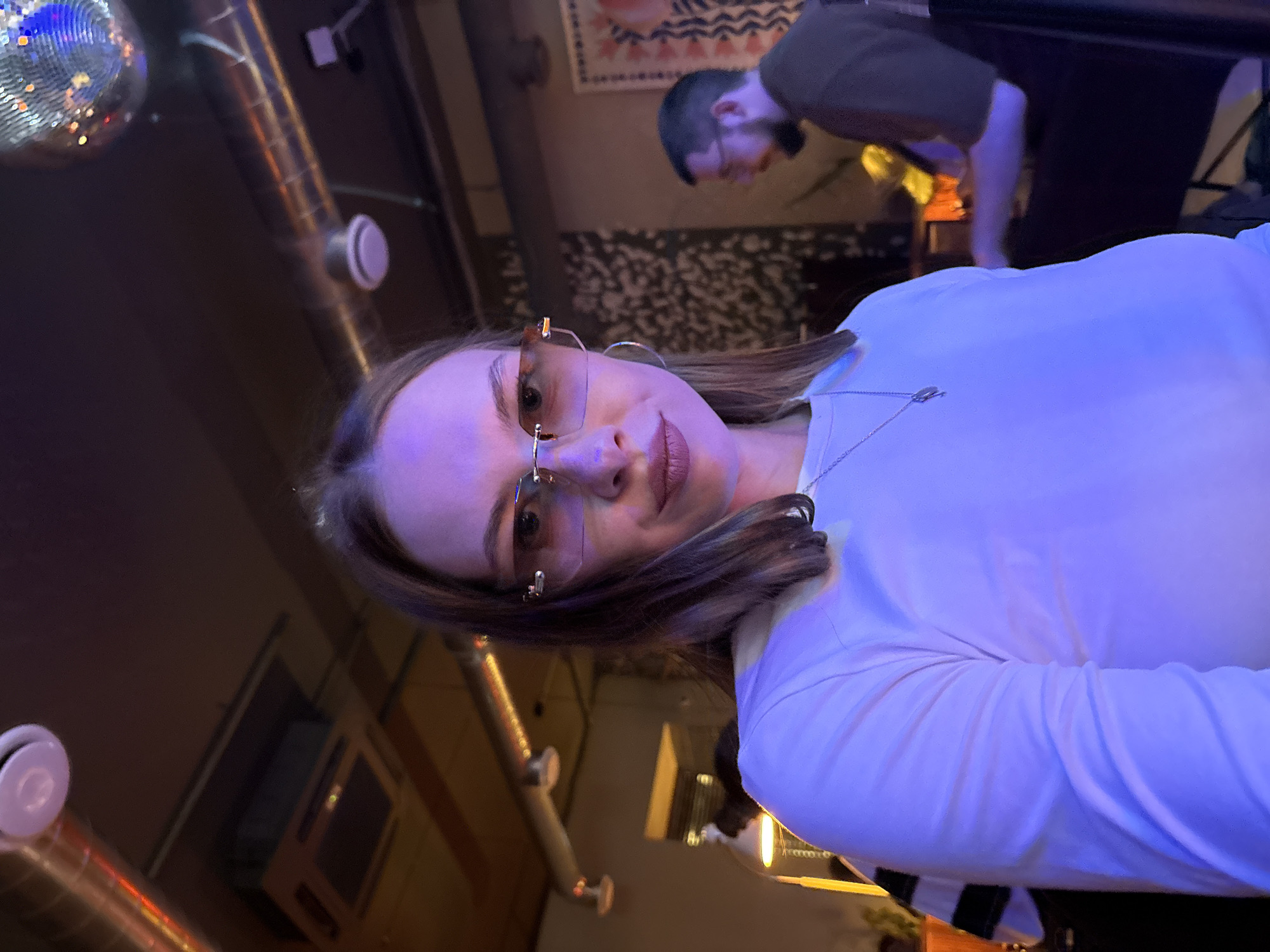 A young woman with brown hair is wearing a white long-sleeved shirt and rimless sunglasses with rose-tinted lenses. She is looking at the camera with a slight smile. In the background, a man with a beard and glasses is seated at a table. The environment appears to be a dimly lit bar or club, with a disco ball visible in the upper right corner and exposed ceiling pipes. The lighting is predominantly purple and orange, creating a warm and inviting atmosphere. There is a decorative tapestry on the wall behind the man. This image was taken in Perm, Russia.