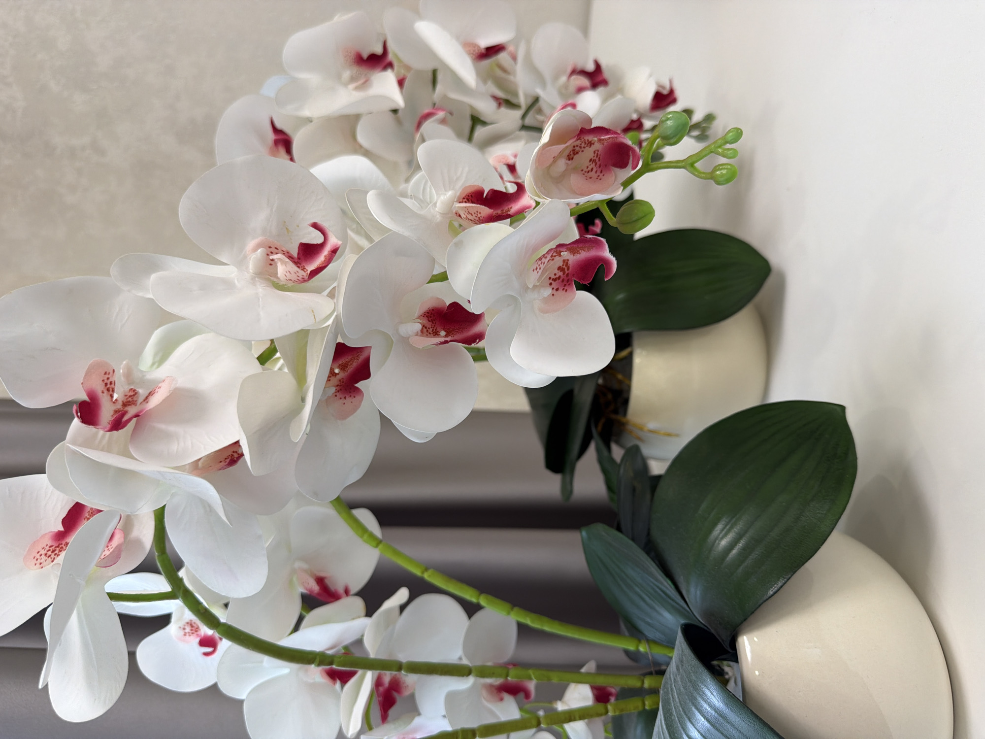 This is a close-up, eye-level, medium shot of artificial white orchids with pink accents. The orchids are in a beige ceramic pot in the foreground, and a smaller beige pot with dark green leaves is visible behind it. The background consists of a grey curtain. The scene is indoors, with a white surface in the foreground. There are no people or animals present.