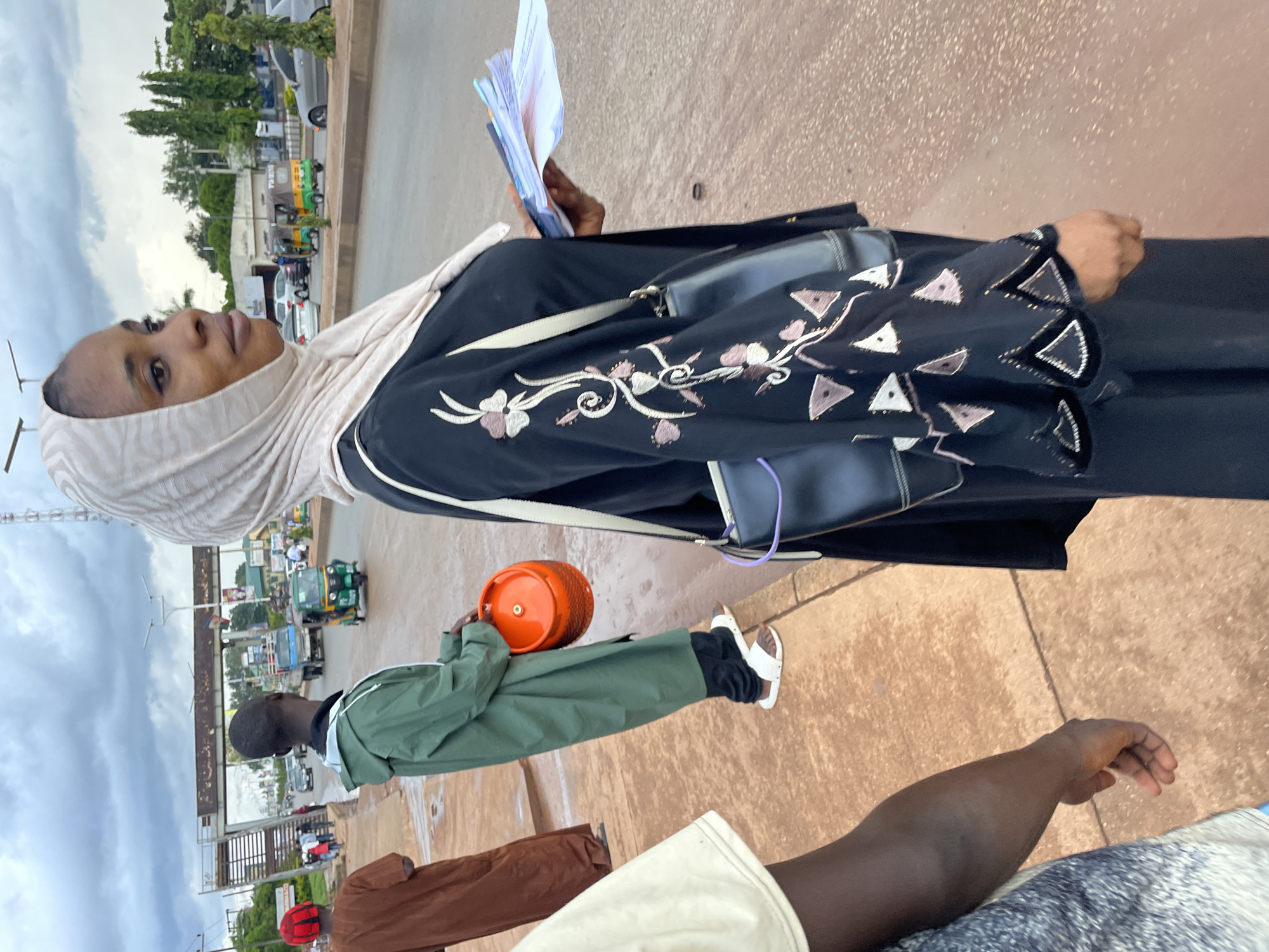 In Jos, Nigeria, on an overcast day, a woman wearing a hijab and a black abaya with embroidered sleeves stands on a street. She is holding a stack of papers and a purse is slung over her shoulder. Next to her, a person in a green raincoat carries a bright orange gas cylinder. In the background, vehicles, including a truck and a tricycle, travel on the wet asphalt road. A pedestrian bridge crosses over the street. Other individuals are visible on the sidewalk and in the distance.