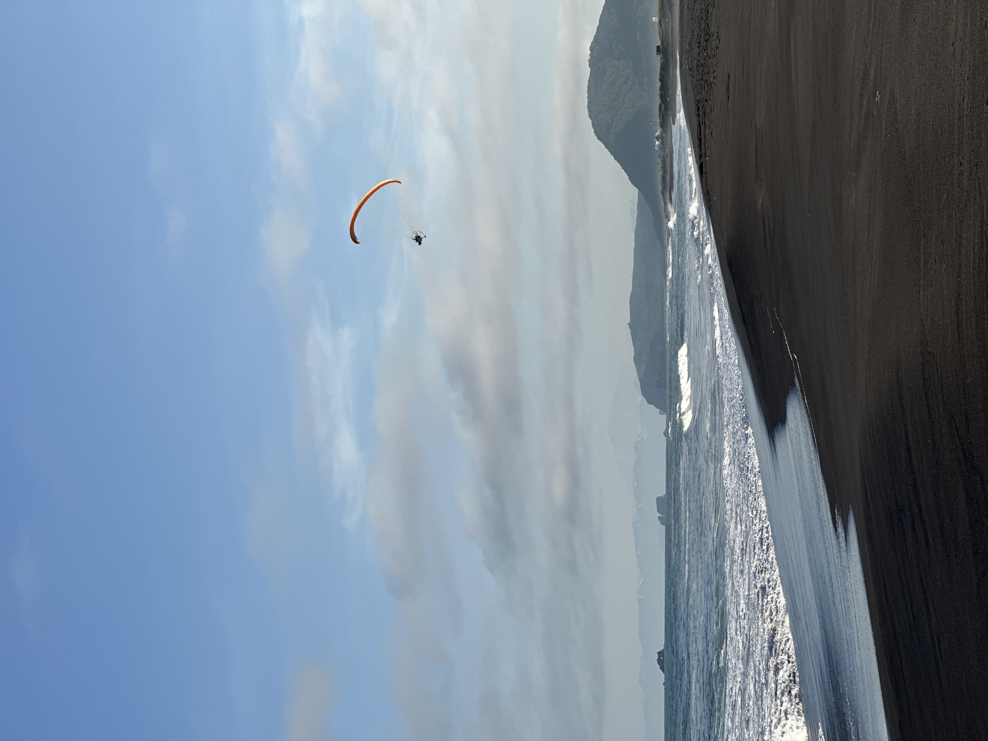 A paramotorist flies under a partly cloudy sky above a black sand beach in Petropavlovsk-Kamchatsky, Russia. The paramotor, orange in color, is a small aircraft with a propeller-driven fan and a wing. It is seen in the upper right quadrant of the image. The beach is dark and appears to be made of volcanic sand. The ocean is a deep blue and the waves are breaking on the shore. In the distance, there are mountains and a few buildings. The sky is a pale blue with scattered white clouds. The overall impression is one of a vast and wild landscape.