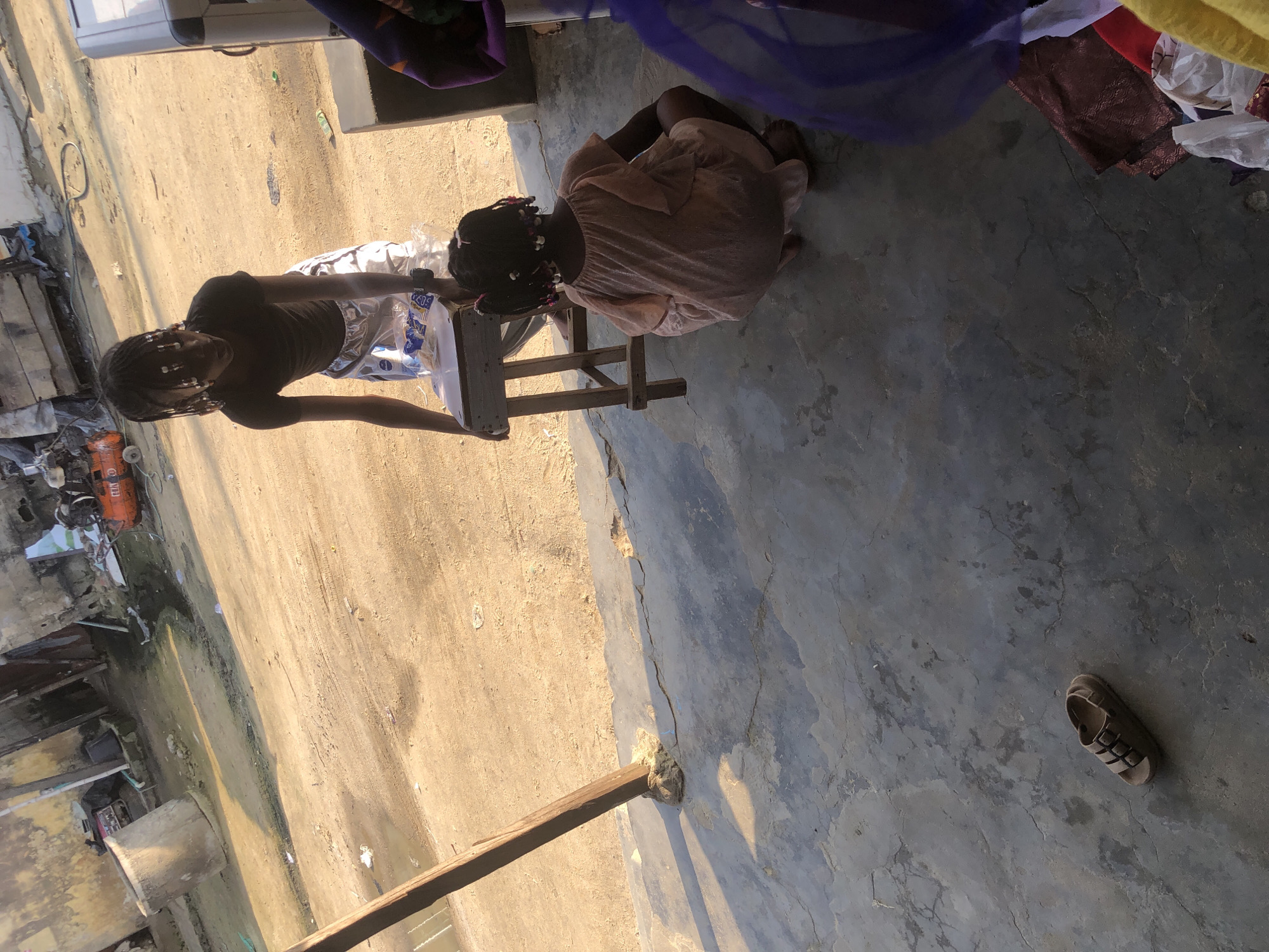 In Ado, Nigeria, an older girl stands next to a wooden stool, offering items from it to a younger girl who is crouching on the ground. The older girl is wearing a dark t-shirt and patterned shorts. The younger girl is wearing a light-colored dress. The ground is dirt and concrete. In the background, there are dilapidated buildings and what appears to be an air compressor. A single sandal lies on the concrete in the foreground. The scene suggests a casual, everyday moment in an outdoor urban environment.