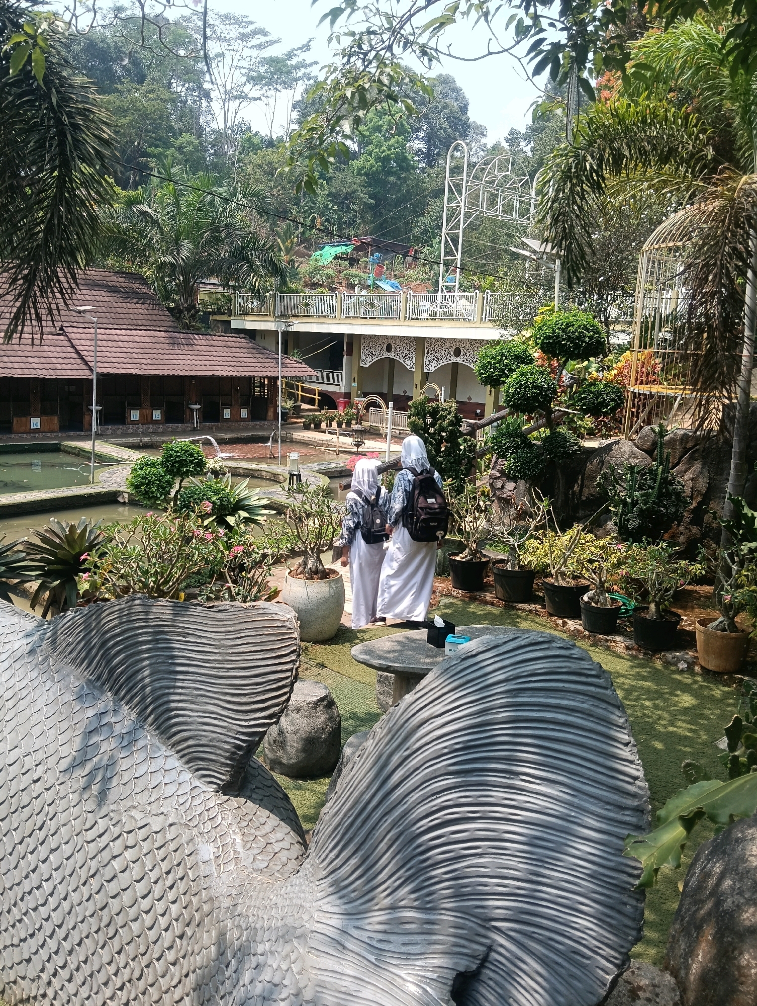 The image depicts an outdoor scene within a park-like setting, possibly a tourist attraction, located in the city of Sempu, Indonesia. In the foreground, two large, stylized grey sculptures resembling fish scales dominate the lower portion of the frame. Two individuals, appearing to be young women wearing long white garments and headscarves, are walking away from the camera towards the right, each carrying a backpack. They are positioned in a garden area with lush greenery, potted plants, and a pond. In the mid-ground, a building with a reddish-brown tiled roof and open-air seating areas is visible, adjacent to the pond. Further in the background, a white structure with ornate white railings and intricate architectural details is situated on higher ground, with a Ferris wheel-like structure and other amusement park elements visible behind it. The environment is characterized by abundant trees, indicating a tropical or subtropical climate.