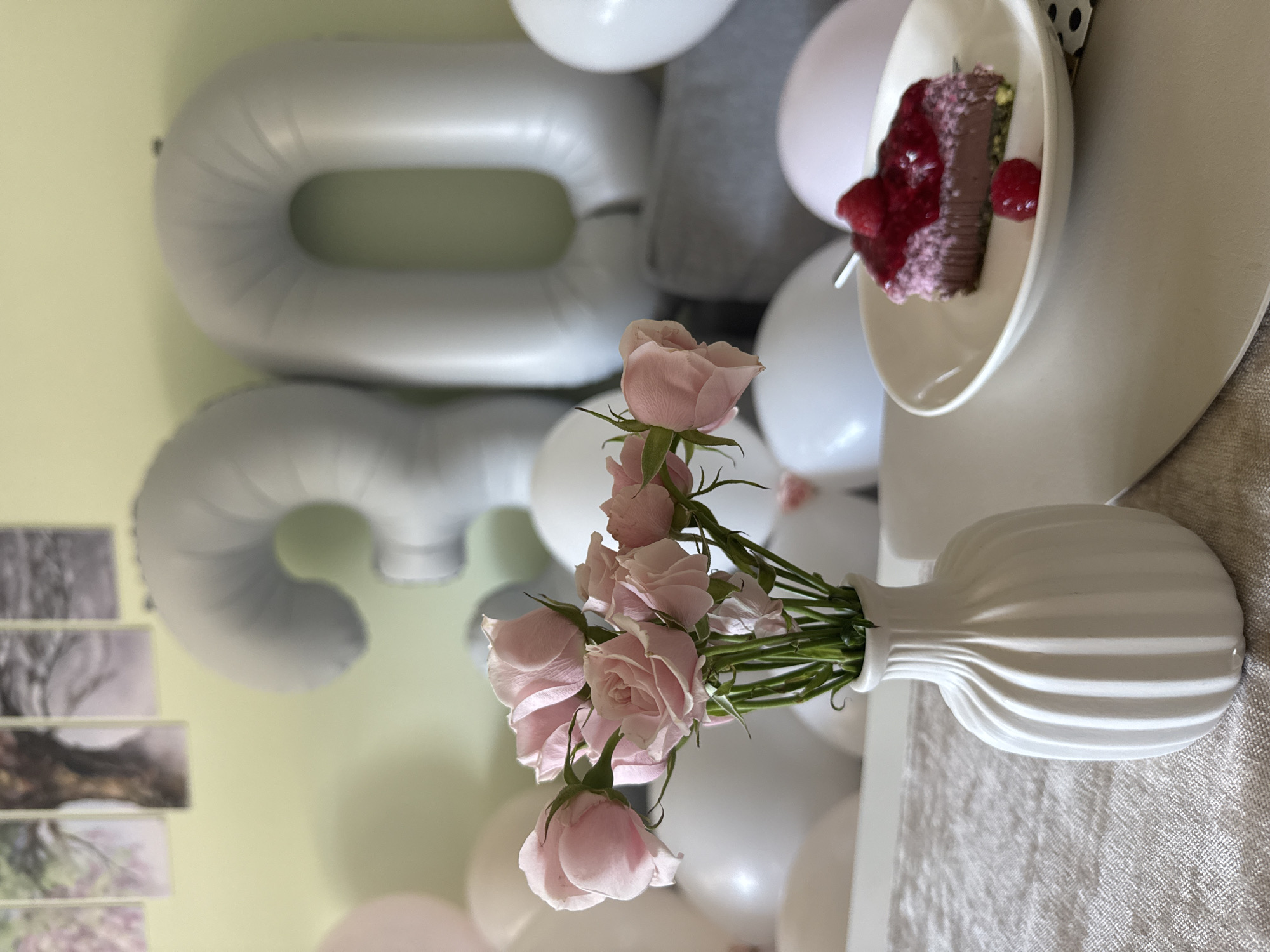The image presents a celebratory scene, possibly a birthday. In the background, large, silver-colored balloon numbers "3" and "0" are partially visible against a pale yellow wall decorated with black and white art. There is a cluster of white and pink balloons, suggesting a festive event. A vase containing pink roses is centrally positioned, its white, ridged design standing out. A slice of chocolate cake adorned with a red fruit topping, likely raspberries or strawberries, is displayed on a small white plate. The scene is likely indoors, with a neutral-colored tablecloth or surface visible beneath the arrangement. The location in Sofia, Bulgaria, is not discernable from the image provided.