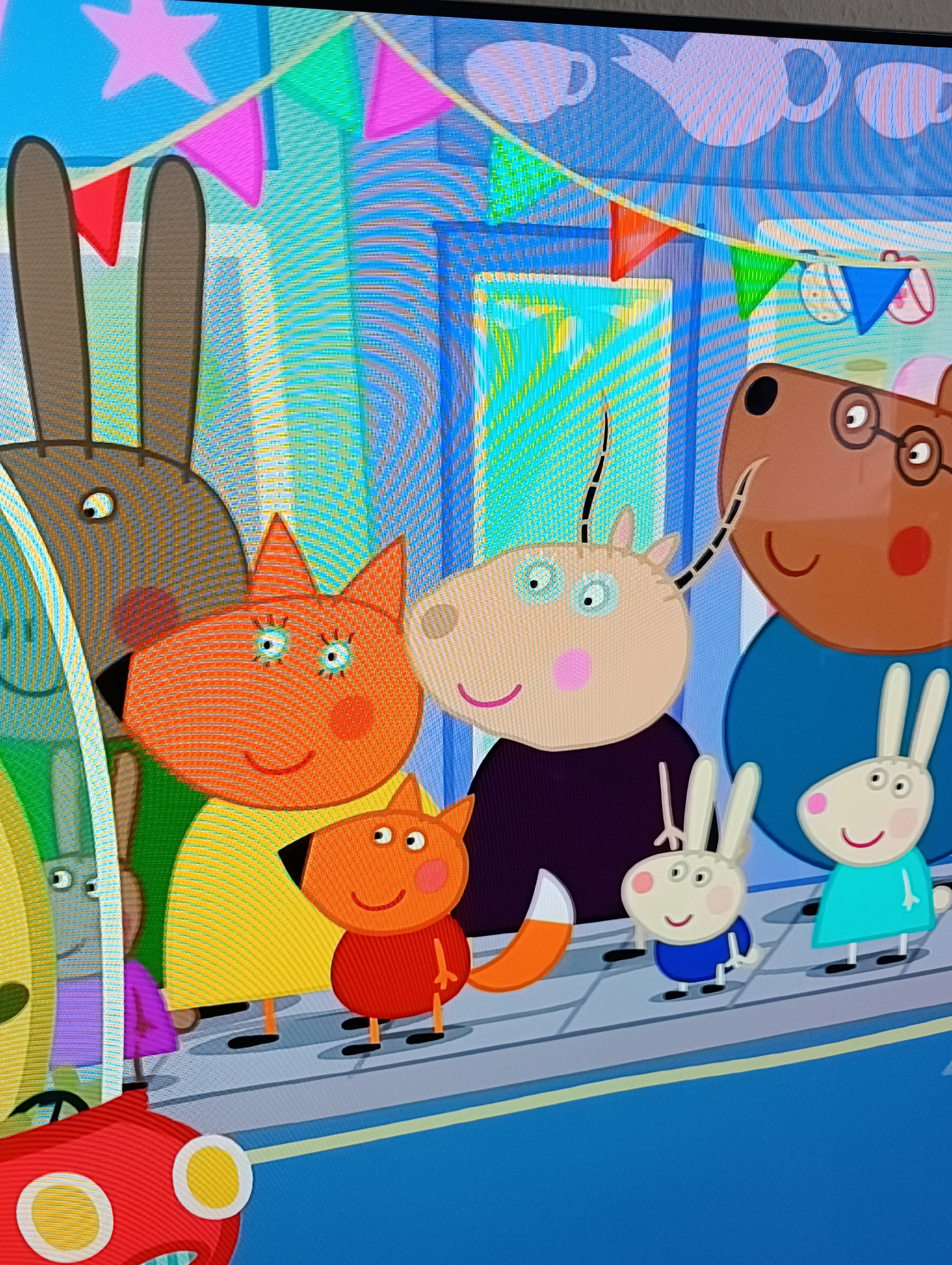 The image shows characters from the children's animated television series "Peppa Pig". The characters appear to be on a sidewalk in a city, possibly participating in a parade or celebration. Bunting decorates the background. The foreground contains cartoon animals including foxes, rabbits, and other species.
