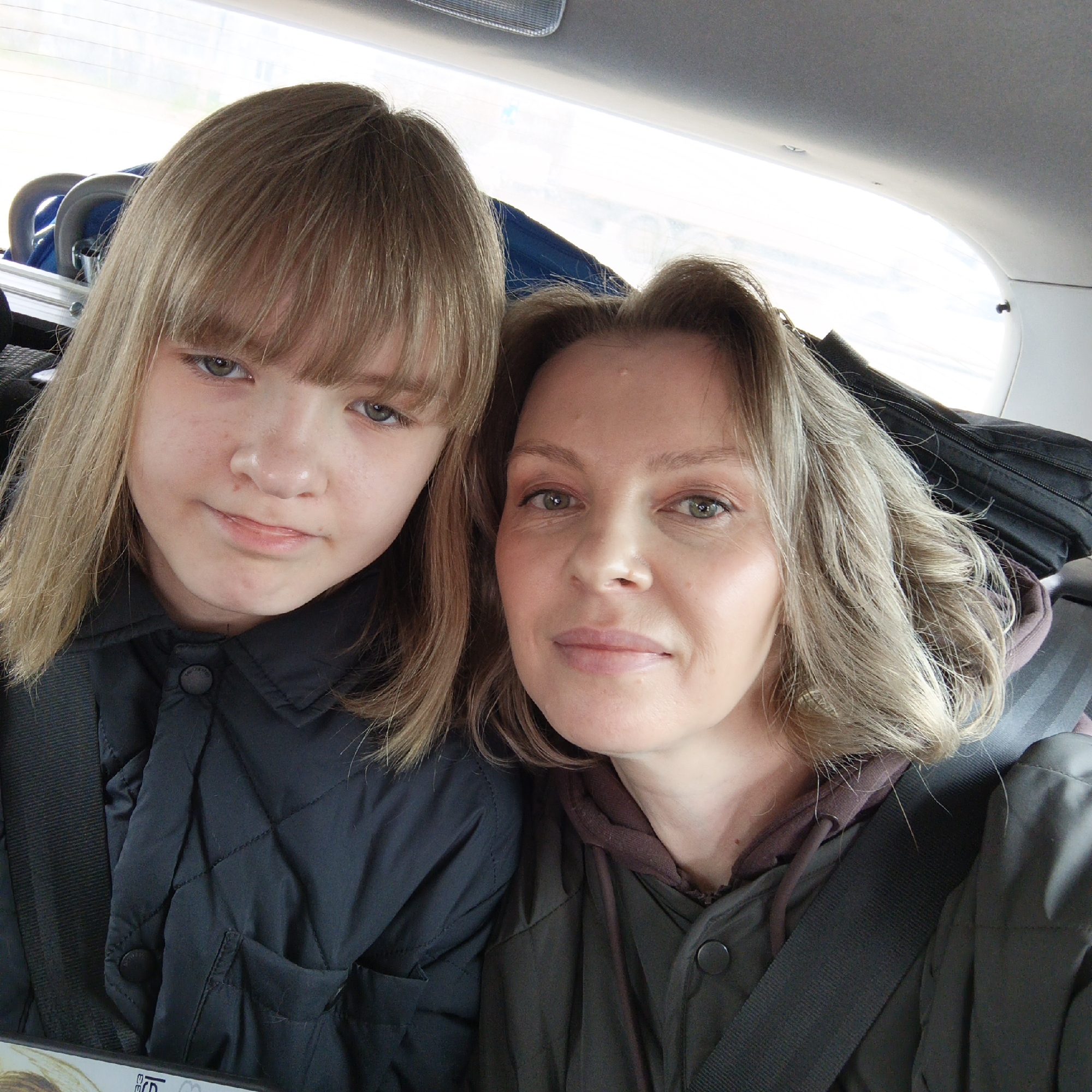 Two people, likely a mother and daughter, are in a car, wearing seatbelts. The mother is on the right and has shoulder-length blonde wavy hair. She is wearing a green jacket with a brown hooded sweatshirt underneath. The daughter is on the left with straight blonde hair with bangs. She is wearing a black quilted jacket. There are items loaded behind them in the car. The location is possibly in Chaikovsky, Russia.