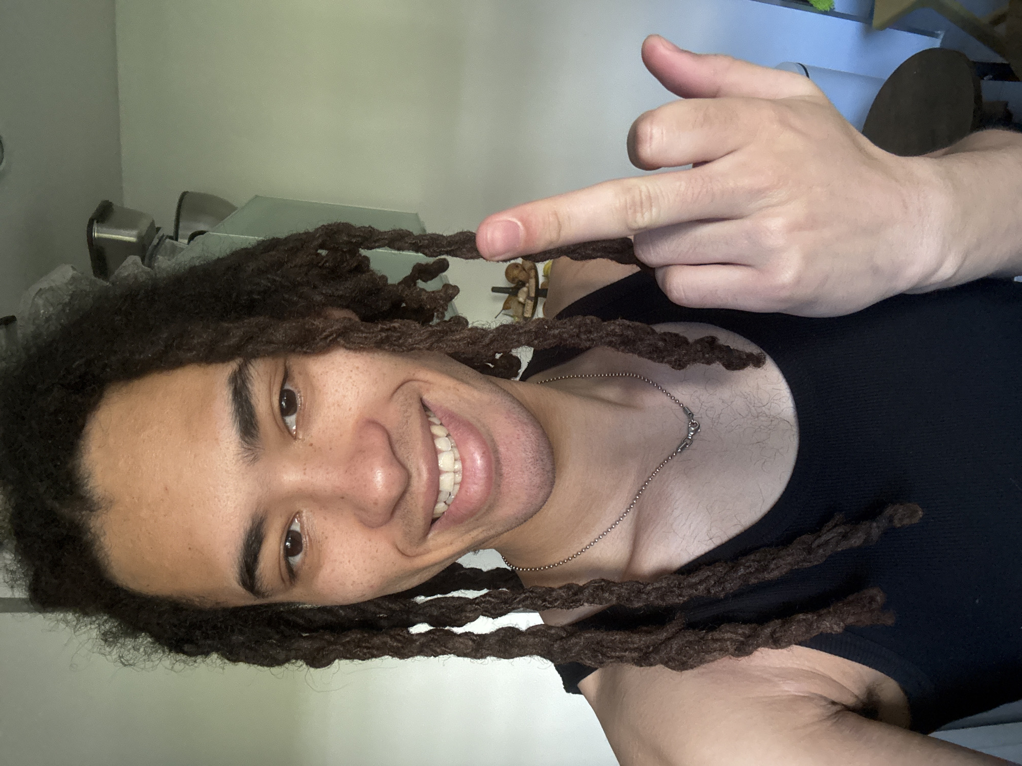 A young adult male is centered in the frame, smiling and facing the camera. He has brown skin, dark curly hair styled in dreadlocks, and brown eyes. He is wearing a black tank top and a silver chain necklace. His left hand is raised, holding one of the dreadlocks between his thumb and forefinger. The background is a plain, light-colored wall with a few items visible, possibly kitchen appliances and a chair. The photograph was not taken in Les Ulis, France.