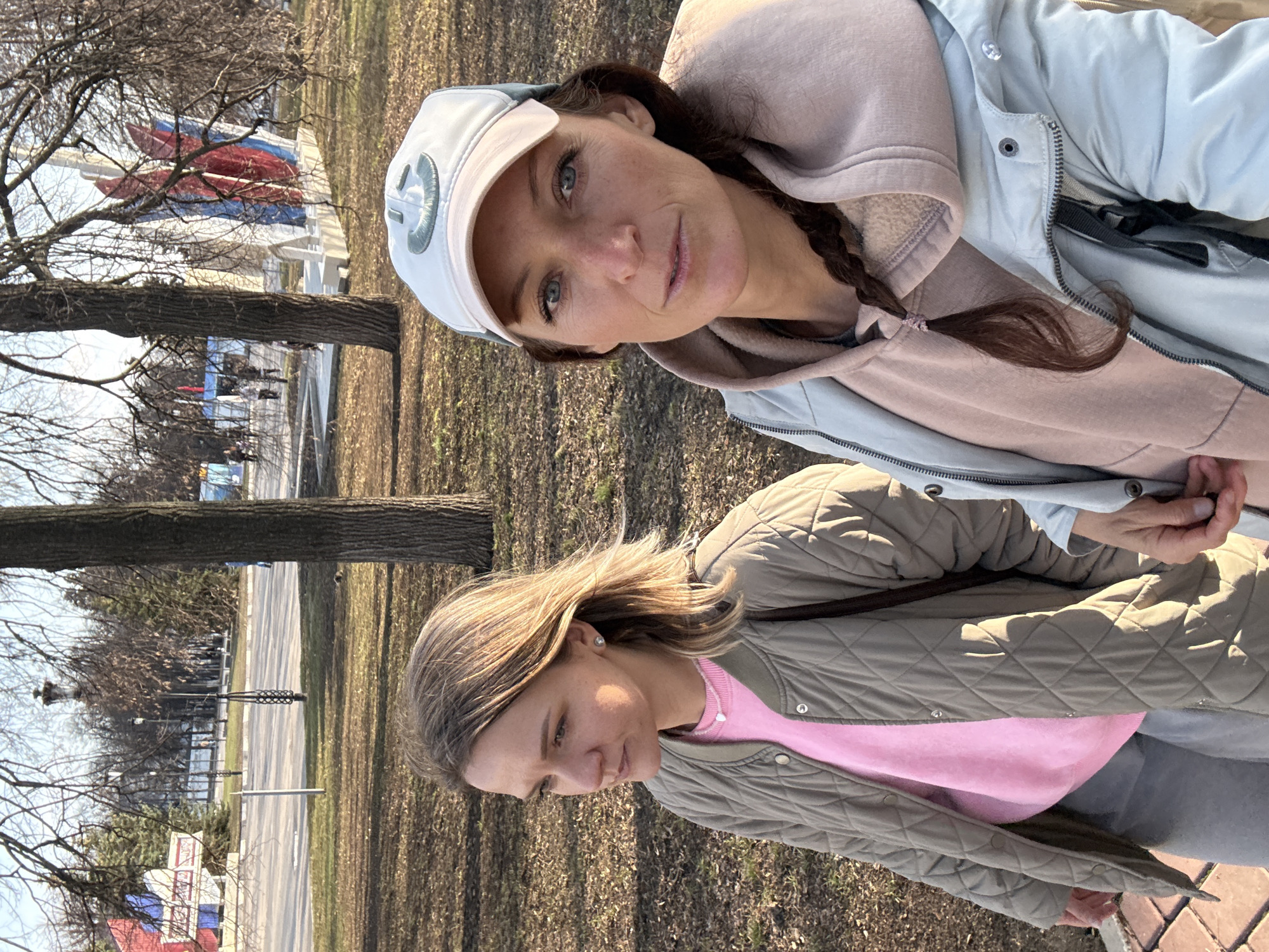 Two women stand outdoors. The woman at the top wears a white baseball cap, a light blue jacket over a light pink hooded sweatshirt, and has her brown hair in a braid. The woman below has blonde hair and wears a pink top under a quilted olive-green jacket. They are standing on a patch of dry grass and fallen leaves. In the background, a road runs between bare trees. Further back, the Russian flag is visible in front of white buildings. This is likely in Ulyanovsk, Russia.
