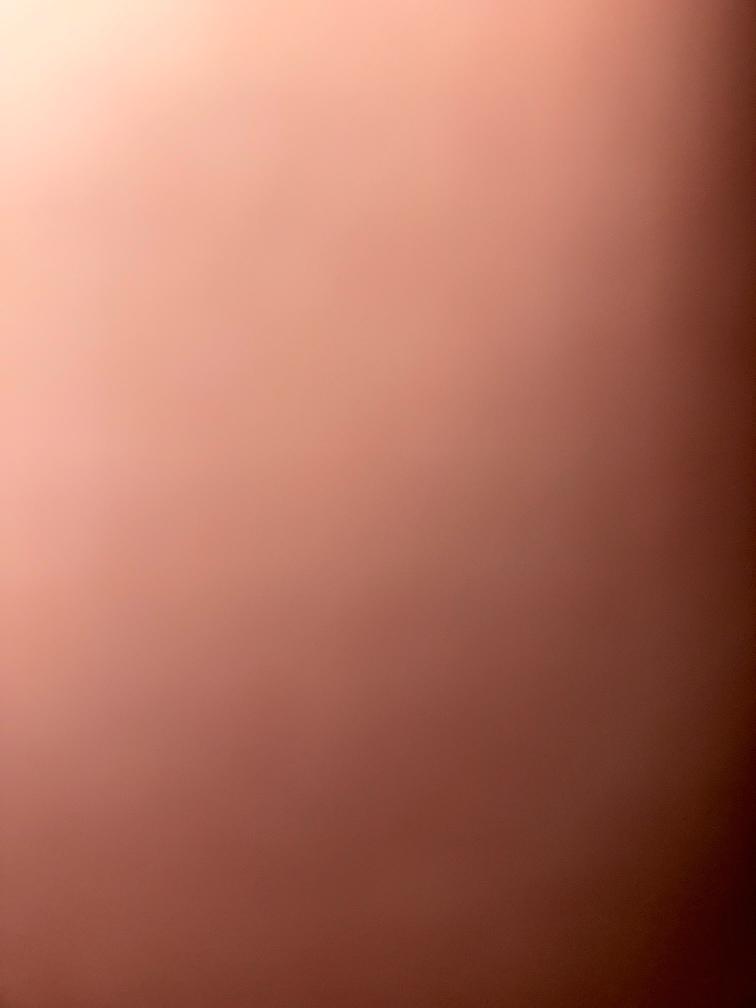 The image is a plain, out-of-focus photograph of a surface that appears to be a wall or a large flat object. The color is a gradient from a peachy-pink on the left side to a darker reddish-brown on the right side. There are no discernible objects, people, or actions depicted in the image. The lighting suggests a light source is present, casting a shadow or creating a gradient effect on the surface. The image is devoid of any context that would place it in Zavolzhye, Russia, or any other specific location.