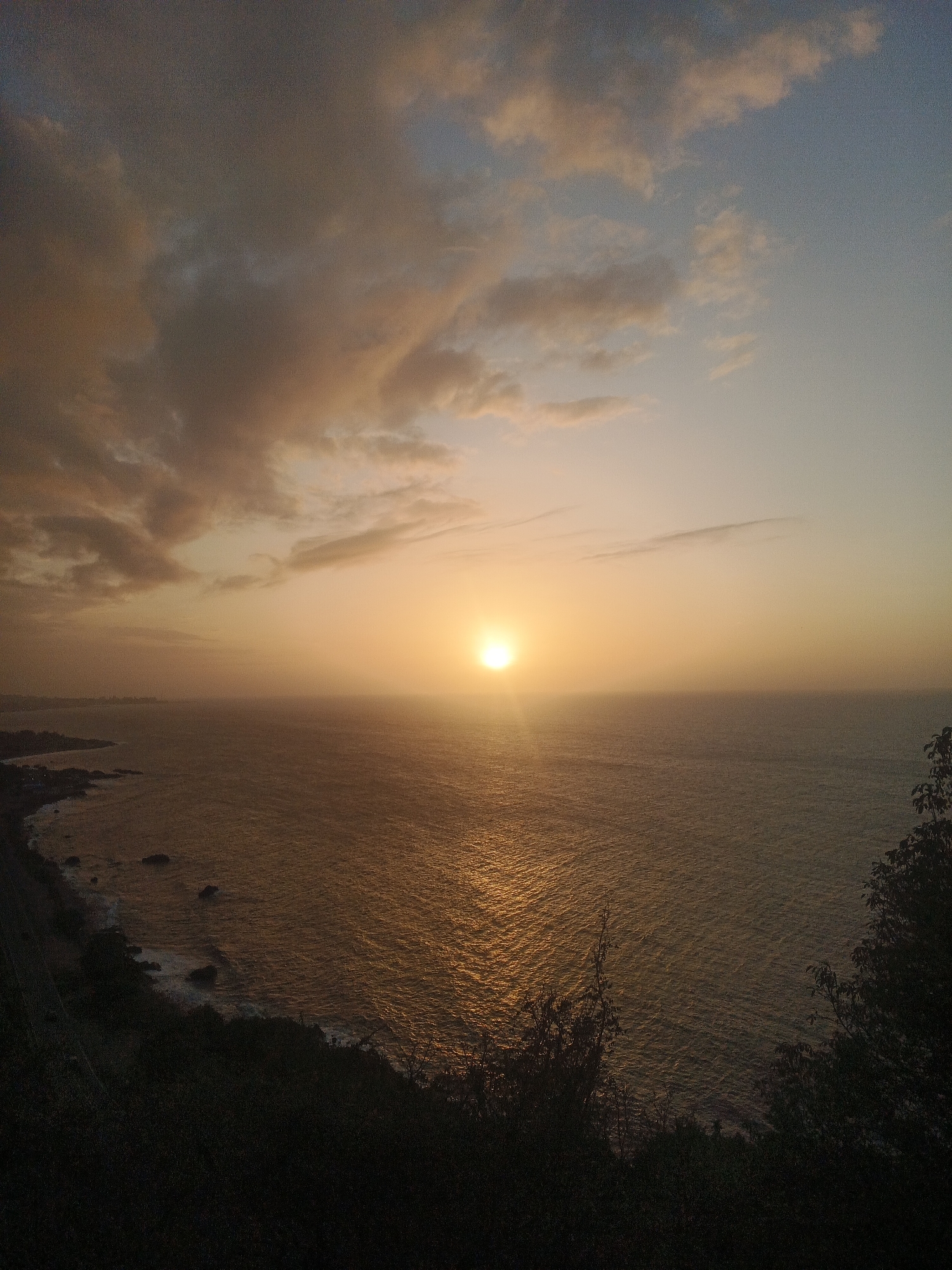 This is a high-angle, wide shot photograph capturing a sunset over the ocean near El Tigrillo, Venezuela. The sun, a bright orb, is positioned in the center-right of the frame, casting a golden reflection across the water. The sky is a gradient of soft blues and oranges, with scattered clouds tinged with pink and yellow hues. In the distance, along the coastline, a town is visible, with lights twinkling as dusk settles. The foreground is dominated by dark, silhouetted trees and foliage, creating a stark contrast with the illuminated seascape. The ocean appears calm, with gentle waves lapping at the shore. The scene evokes a sense of tranquility and natural beauty.