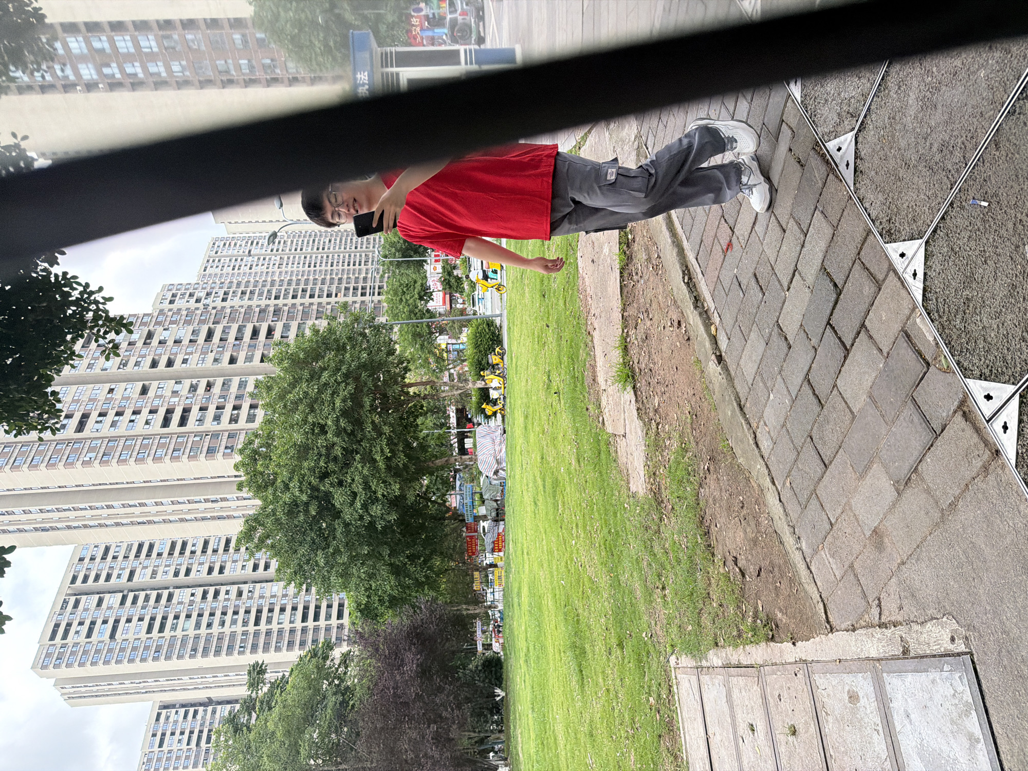 The image shows an individual wearing a red t-shirt, grey pants, and white shoes standing on a brick sidewalk next to a grassy area with trees in Chongqing High-tech Zone, China. The person is holding a smartphone and appears to be taking a selfie. In the background, there are tall apartment buildings and trees. Yellow shared bikes are visible in the distance. The image appears to be taken from inside a vehicle, looking out of the window.