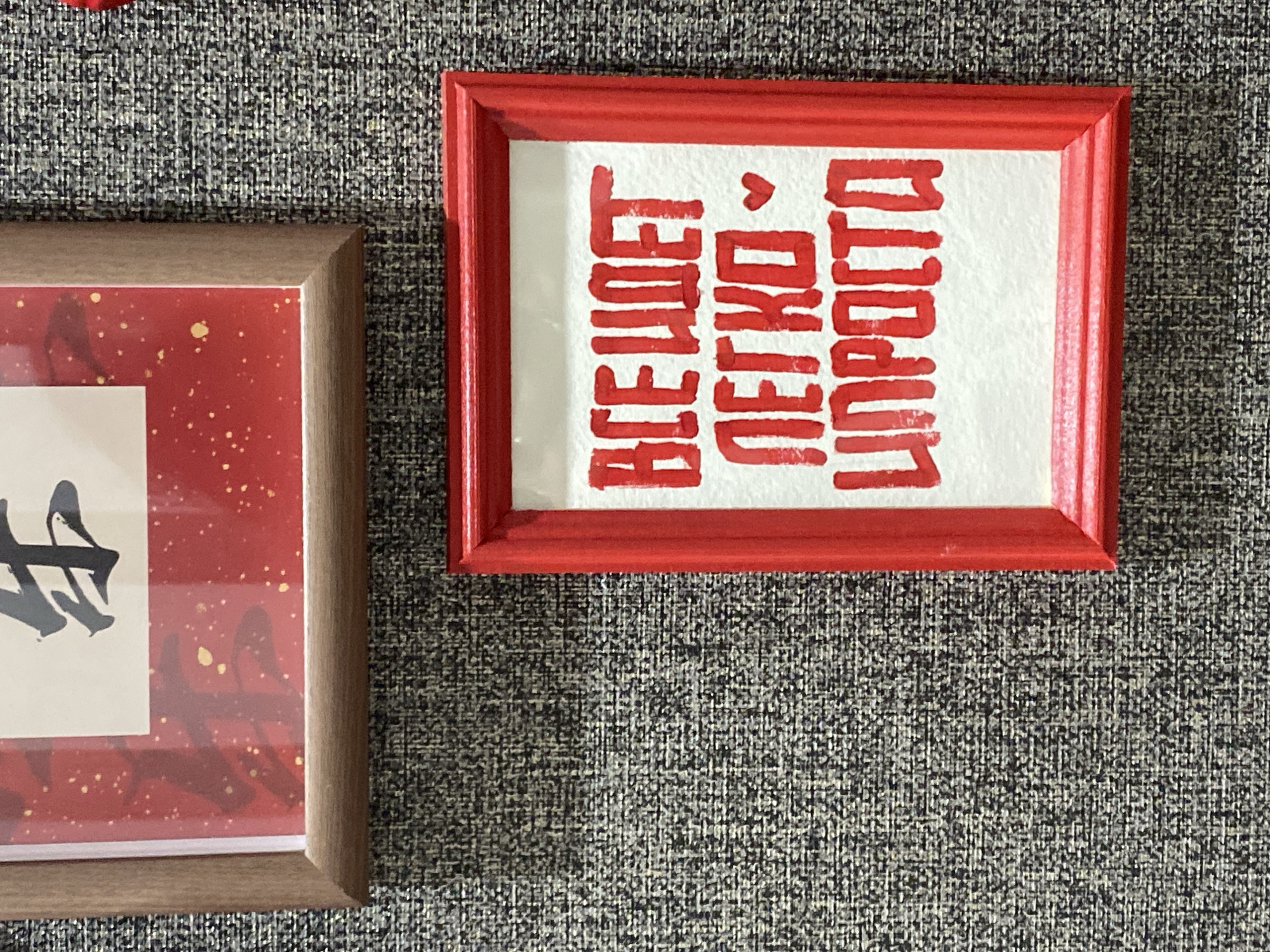 The image shows two framed pieces of art hanging on a textured gray wall. The top frame is a wooden frame with a reddish-brown hue, containing a piece of art with red and gold accents and black calligraphy on a white background. The bottom frame is a bright red frame, enclosing a white mat with red lettering in a Cyrillic script. The red lettering on the bottom piece reads "ВСЕ ИДЕТ ЛЕГКО ♥ ПРОСТО" which translates to "Everything goes easily ♥ Simply". The overall scene appears to be an indoor setting, possibly a home or a gallery, with no visible urban features or specific landmarks to indicate a location within the city of Chaikovsky, Russia.