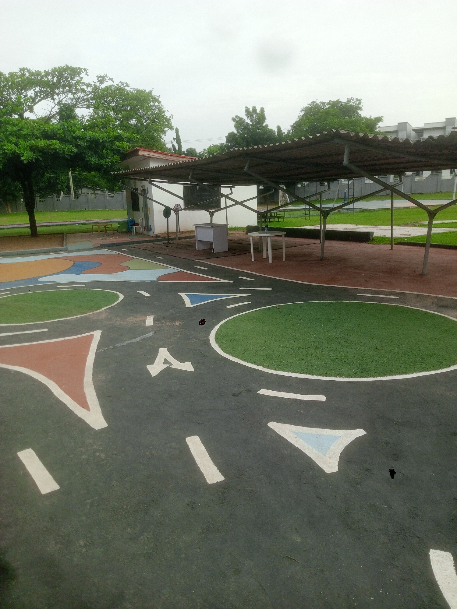 The image depicts a playground area in Abuja, Nigeria. The foreground features a painted road network on a dark asphalt surface, complete with white lane markings, colorful triangular and circular zones, and an "A" symbol. A covered shelter supported by metal beams stands in the background, containing a white table. The playground is situated in an outdoor area with green trees and grass, under an overcast sky. There are no people visible in the frame.