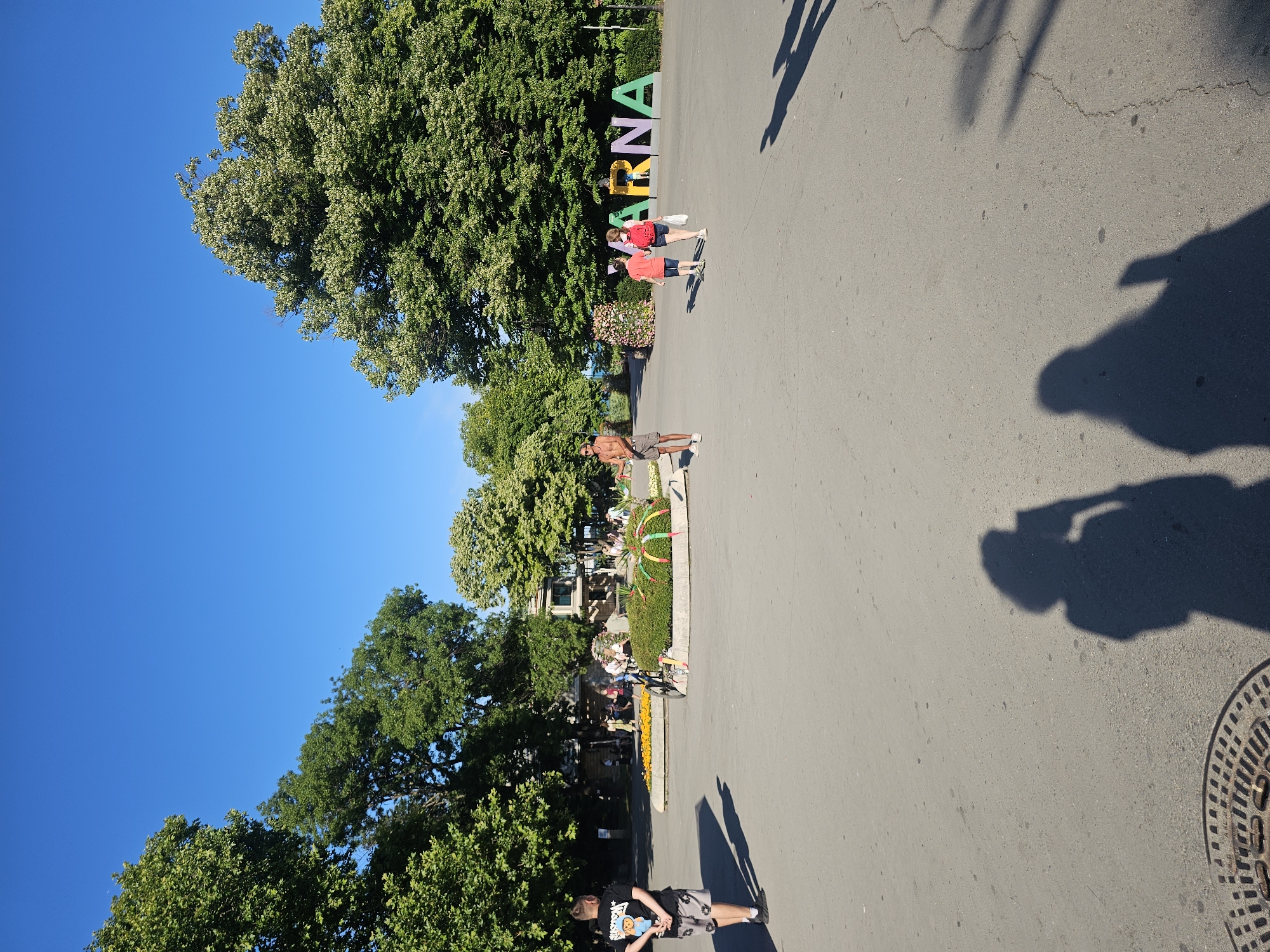 A bright, sunny day unfolds in an urban park. A person wearing a black t-shirt with a teddy bear graphic and gray shorts stands facing away from the camera. Further into the park, a man with shorts and no shirt walks with a cell phone in his hand. Behind him, two children, one wearing a red shirt and the other a pink shirt, walk together, the one in red carrying a white bag. Large, leafy trees dominate the background, casting shadows on the paved ground. In the distance, large, colorful letters spell out "VARNA." People are scattered throughout the park, some seated and others standing. The scene suggests a leisurely day in a public green space.