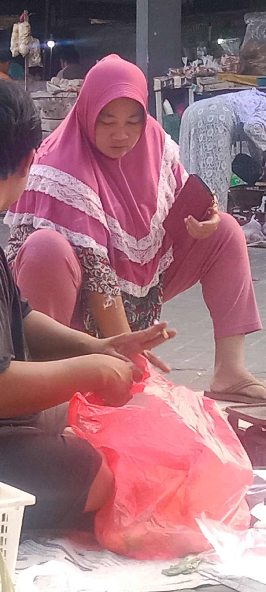 This is a full-frame, eye-level, outdoor photograph of two individuals, seemingly engaged in a transaction at a market stall. The primary subject, a person wearing a pink hijab and a matching pink tunic with white lace trim, is crouched down, facing away from the camera, and looking at their hands which are manipulating a large, crumpled, bright pink plastic bag filled with items. Their right hand is also holding a dark red wallet or clutch. The secondary subject, seen from the shoulder down on the left, is also crouched and appears to be handing something to the primary subject. This individual is wearing a dark t-shirt and has their hands near the pink plastic bag. In the background, blurred figures and market stalls suggest a bustling environment. The ground surface is paved. The overall impression is a snapshot of daily life at a local Indonesian market, possibly in Kras, based on the provided context.