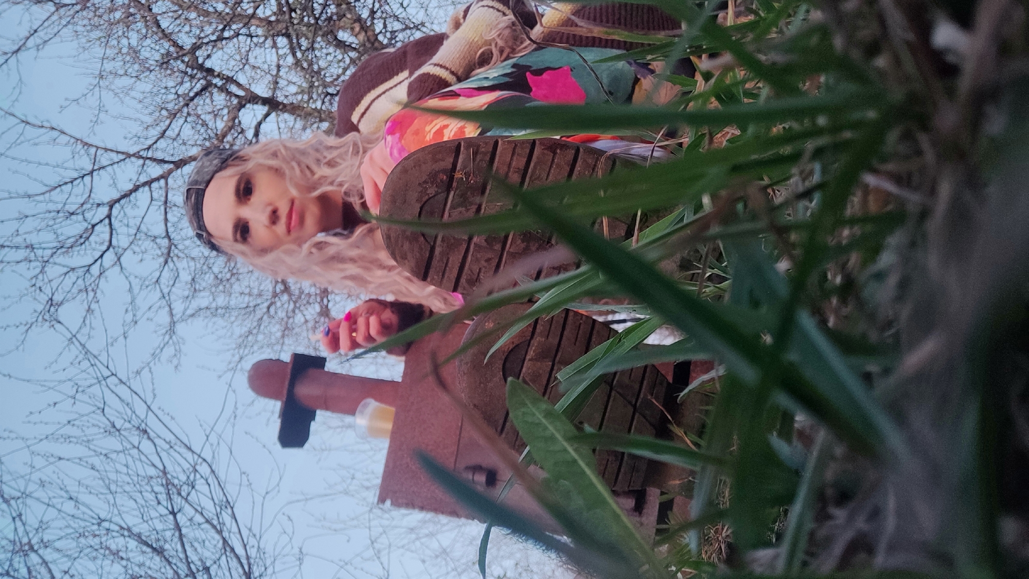 A woman with blonde curly hair, wearing a camouflage cap and a colorful jacket, stands in an outdoor setting in the city of Chaikovsky, Russia. She is positioned behind what appears to be a worn, weathered metal structure, possibly a piece of equipment or part of a building's remains. Tall grass partially obscures the foreground and the bottom of her boots are visible. A backdrop of bare tree branches against a pale sky suggests it might be late autumn or early spring. The scene is shot from a low angle, looking upward towards the woman. She is looking down towards the camera.