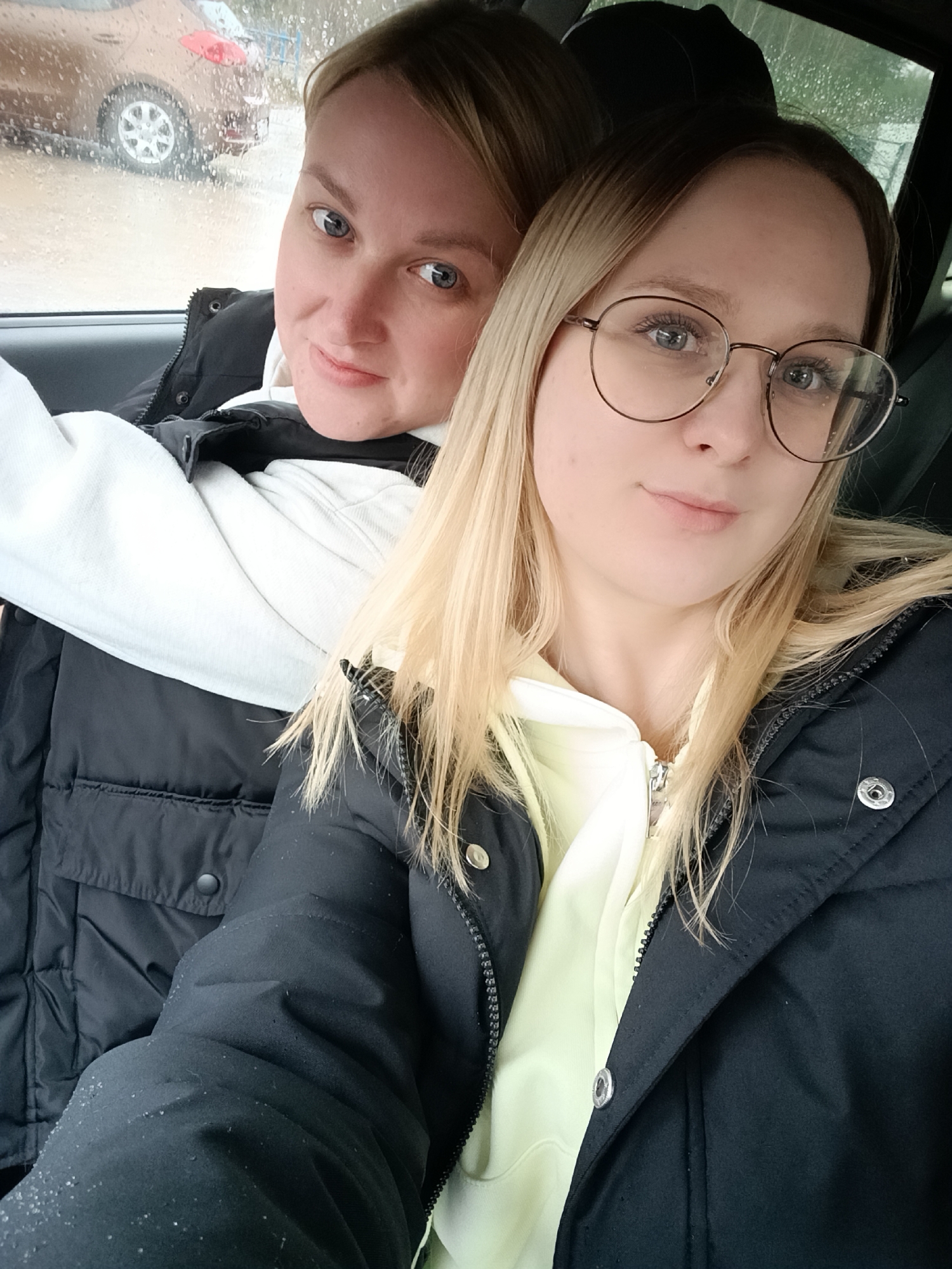 This close-up image shows two women inside a car on a rainy day, likely in Noviy, Russia. The woman in the foreground has long blonde hair, wears round metal-framed glasses, a black puffer jacket over a pale yellow hoodie, and has a serene expression with a subtle smile. Small, sparkling particles are visible on her jacket sleeve. Behind her, partially visible and slightly out of focus, is another woman with light blonde hair, blue eyes, and a gentle smile, wearing a black puffer gilet over a white hoodie.
They appear to be sitting on the passenger side of the vehicle. Through the car window, which is covered in raindrops, a brown car and hints of a blue structure or fence are visible in the background, suggesting they are parked in an outdoor area. The diffused lighting and visible raindrops confirm it is an overcast and wet day. Both subjects are looking towards the camera, suggesting a selfie or posed photograph. No discernible text is visible.