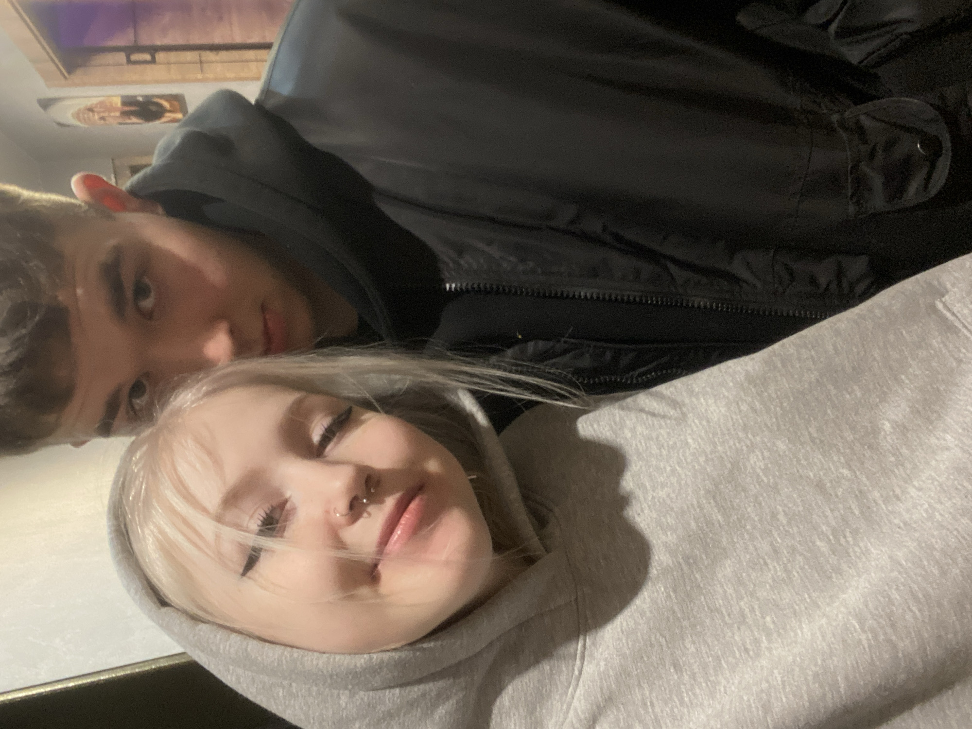 An indoor photograph, rotated diagonally, captures two young individuals, a male and a female, lying down in a relaxed pose in Izhevsk, Russia.
In the foreground, positioned in the lower right portion of the frame, is a female with blonde hair and fair skin. She is wearing a light gray hooded sweatshirt, with the hood pulled up. She has multiple visible piercings on her nose, including a nostril ring and a septum ring. Her eyes are mostly closed, and a subtle, relaxed smile suggests contentment or happiness.
Behind her, situated in the upper left of the frame, is a male with short, dark hair and fair skin. He is wearing a dark, possibly black, jacket with a visible zipper and a small, buttoned pocket detail on the sleeve. He looks directly at the camera with a neutral expression.
The background is slightly out of focus, showing a wall with what appears to be framed artwork or pictures. The lighting suggests an indoor setting, possibly under artificial light or during the evening, creating some soft shadows, particularly around the female's head on her hoodie. The overall scene conveys a casual, intimate, and relaxed moment, possibly a selfie shared between two people. There is no visible text in the image.