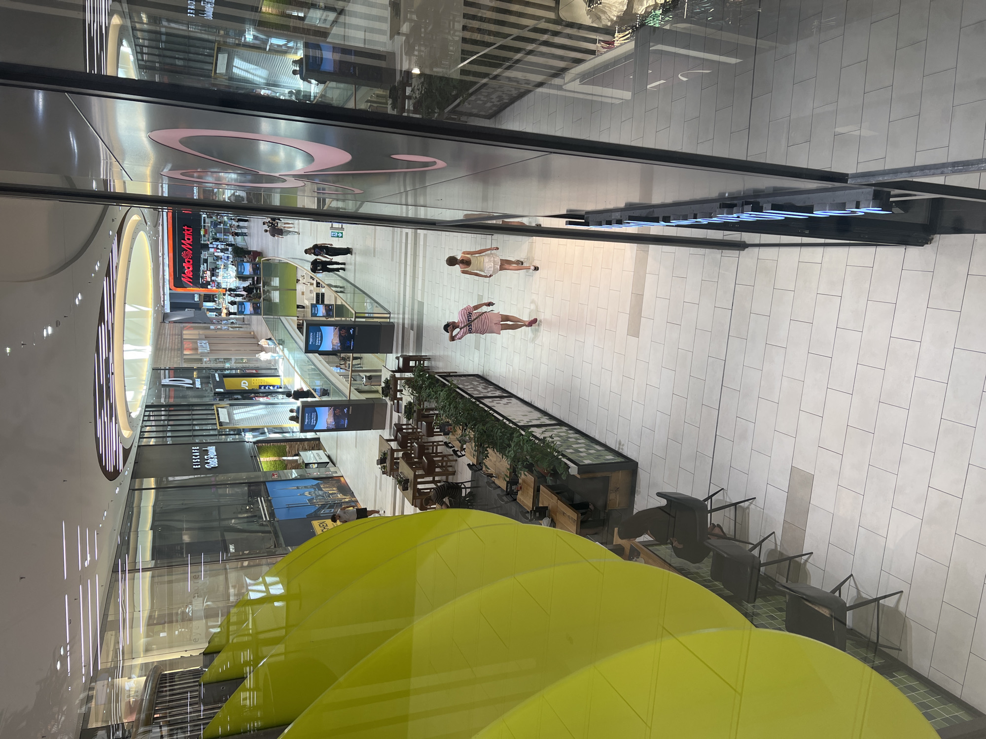 The image shows a brightly lit, modern shopping mall with multiple levels and a high, open ceiling. The flooring is tiled in a light color, and there are numerous shops and businesses visible along the walkways. A large, vibrant yellow sculptural element dominates the left side of the foreground, reflecting the mall's interior.
In the middle ground, two young women are walking away from the camera down a wide walkway. The woman on the left is wearing a pink t-shirt and pink shorts, and the woman on the right is wearing a white tank top and a white skirt. They are walking past a row of tables and chairs set up outside a restaurant or cafe area.
Further back, other shoppers are visible walking and interacting within the mall. Signs for businesses like "MediaMarkt" are prominent. The mall features large glass storefronts, modern lighting fixtures, and decorative greenery along some of the shopfronts. The overall impression is a busy, contemporary retail environment.
The image is identified as being taken in Aachen, Germany.