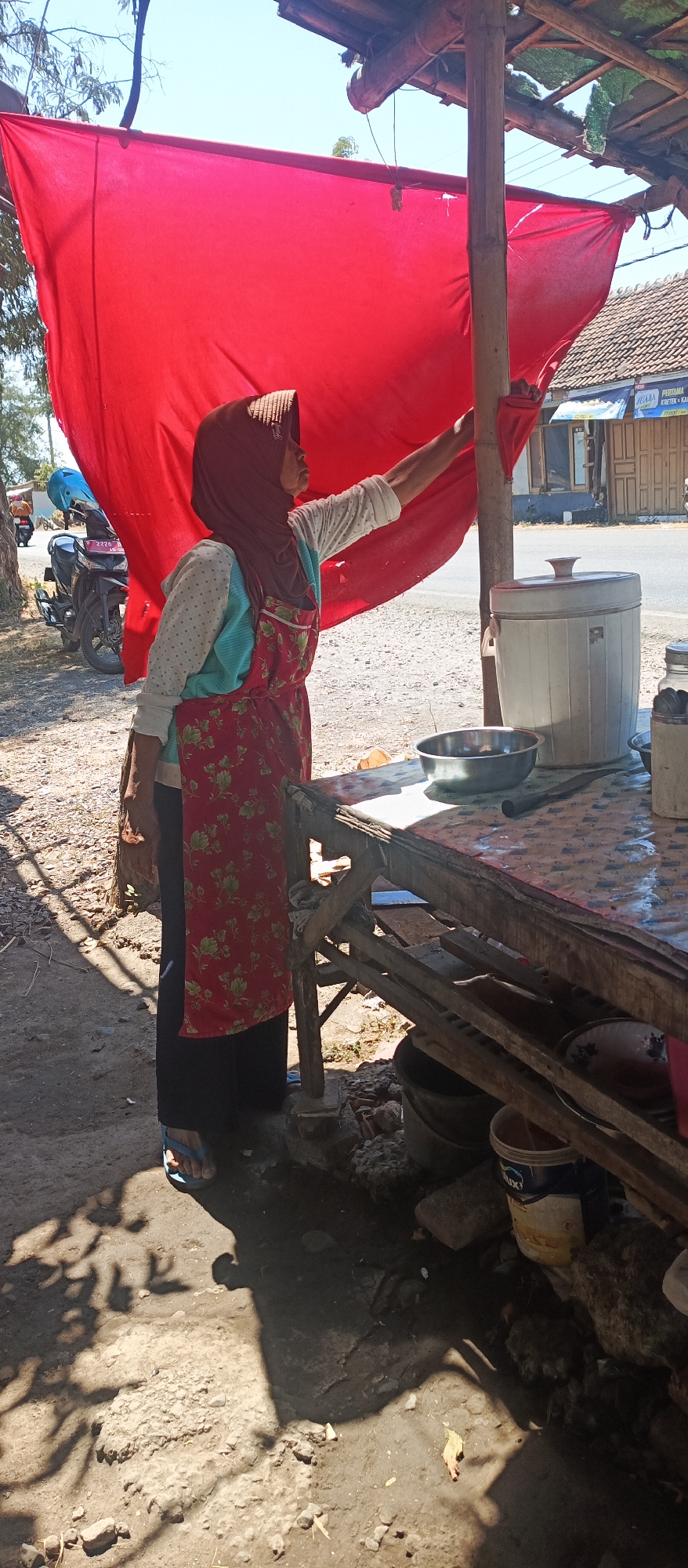 A woman wearing a red floral apron and a brown hijab stands at an outdoor food stall in Jatisari, Indonesia. She is reaching up to adjust a large piece of red fabric that serves as a canopy. In front of her is a wooden table with various items, including a metal bowl, a knife, and a large white container. A motorcycle is parked behind her to the left. The ground is dirt and gravel, and shadows of trees are cast on the ground. In the background, there are buildings and a road. The overall scene suggests a humble street-side business operating in a sunny, outdoor environment.