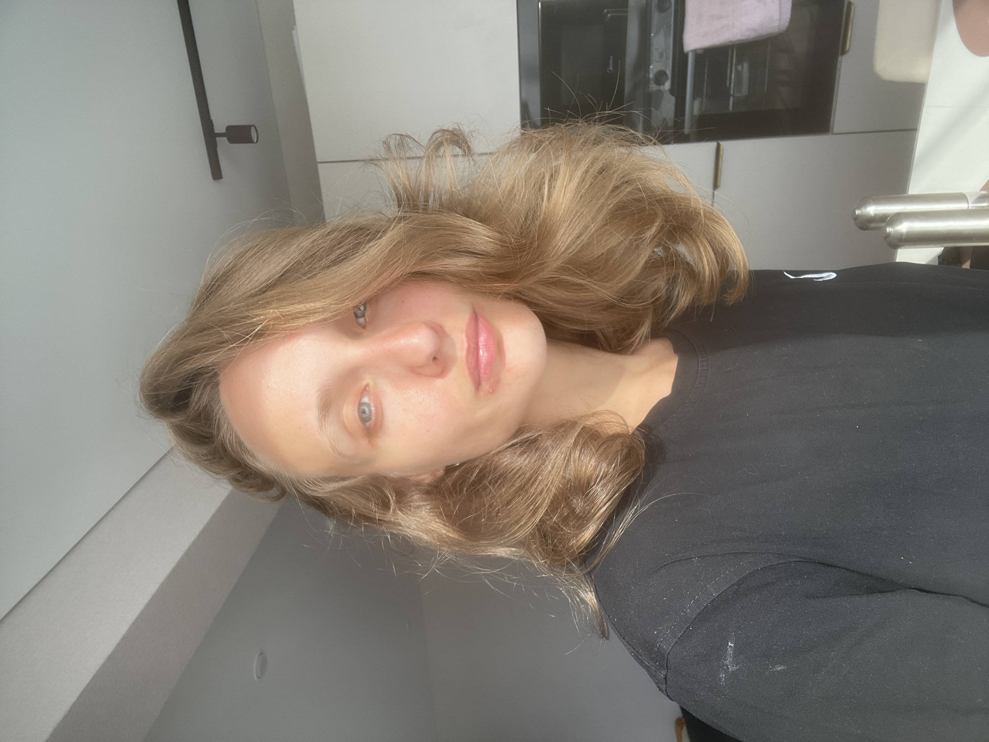 A young Caucasian woman with medium-length blonde hair is posing for a selfie in what appears to be a kitchen. She is wearing a black t-shirt. The background shows kitchen cabinets, an oven, and a light fixture on the ceiling. The lighting suggests it is daytime, with sunlight coming from the left. There are no specific identifiable landmarks that would place this image in Podshivalovo, Russia.