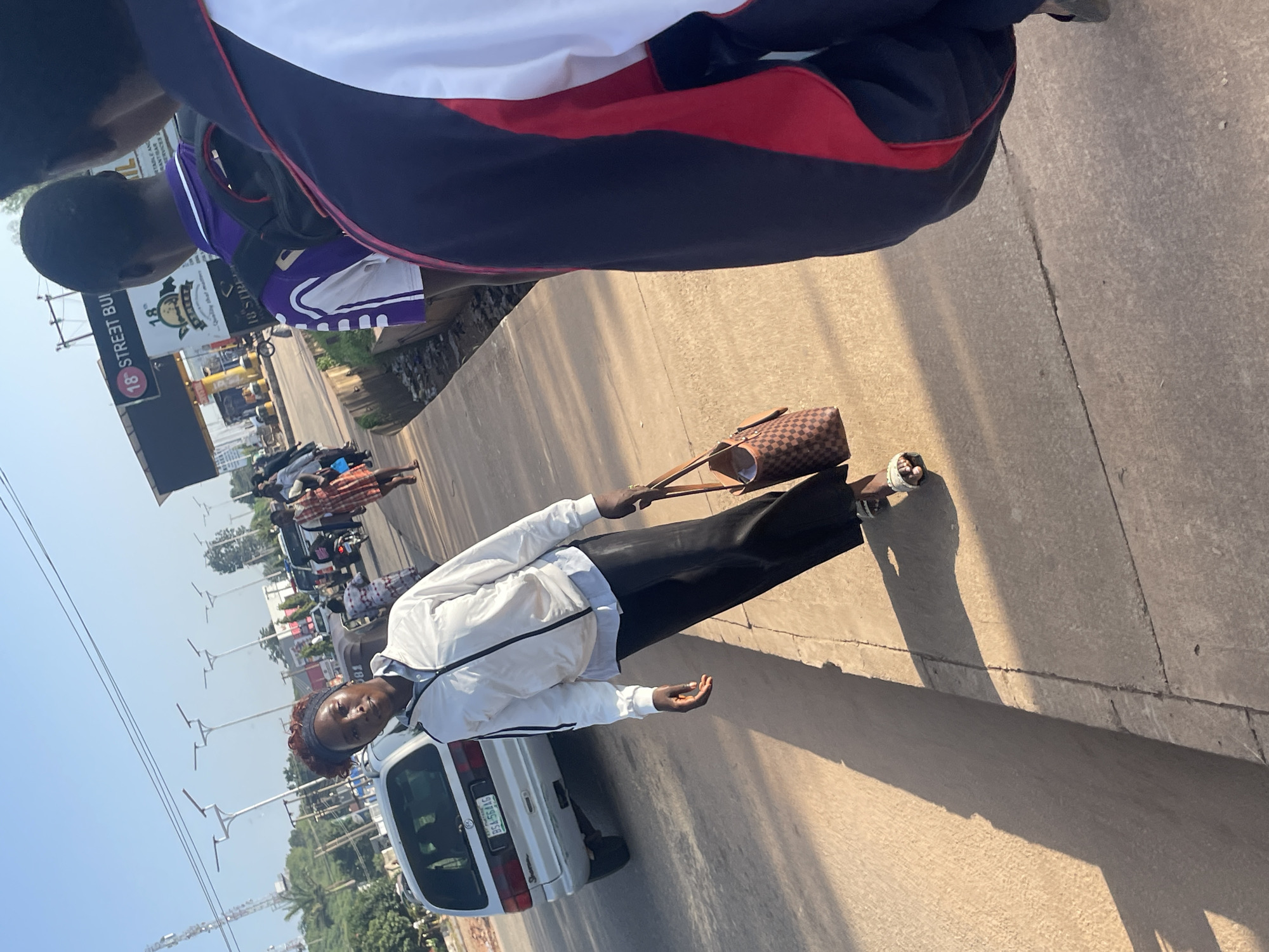 A young woman walks on a sidewalk in the city of Jos, Nigeria. She is wearing a white jacket over a light blue shirt and black pants. She carries a brown patterned handbag in her right hand. In the background, a silver minivan is parked on the street. The sky is clear and blue. The scene is illuminated by bright sunlight, casting long shadows on the sidewalk.