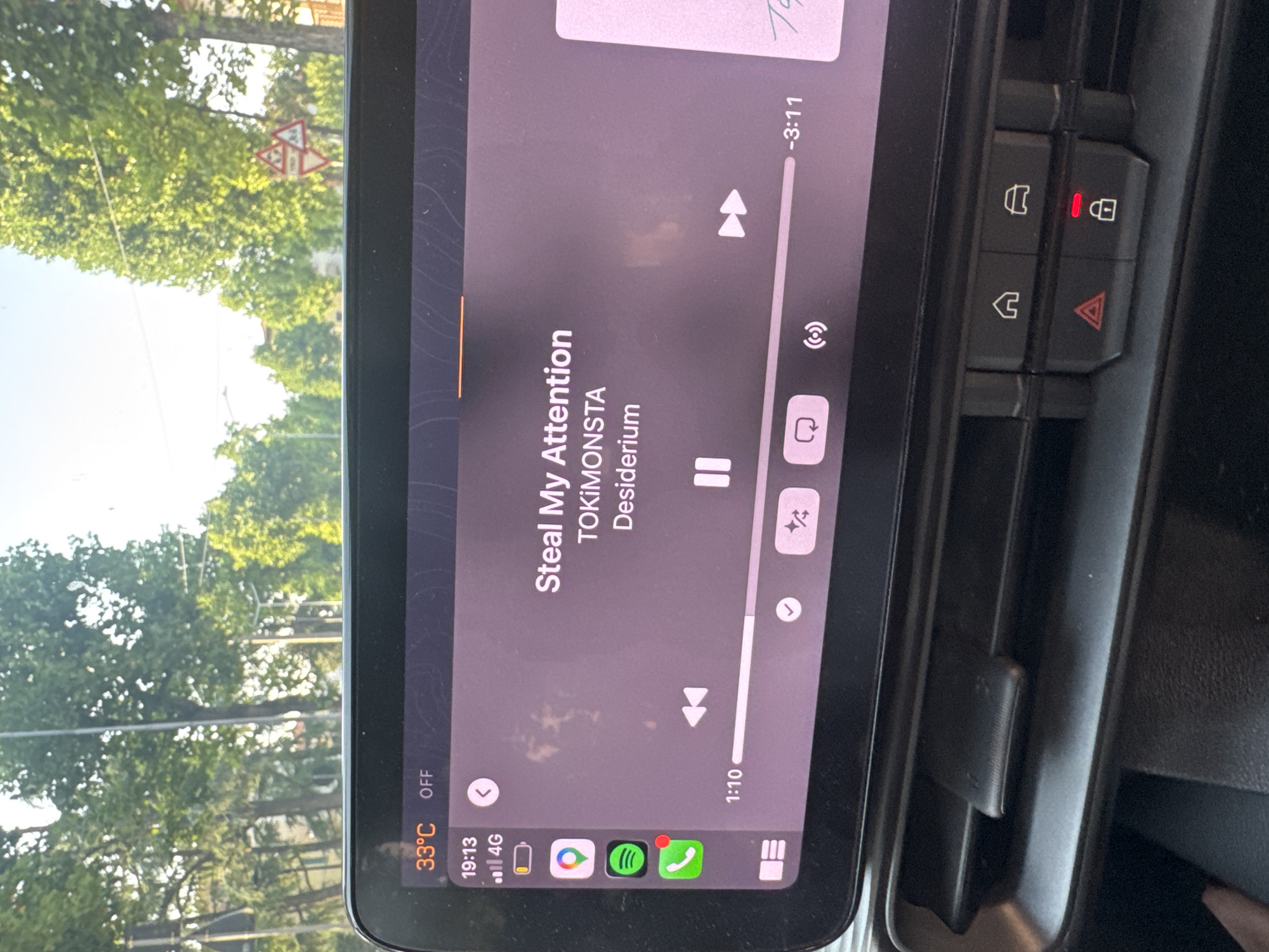 The image shows a car's dashboard display, likely in Chiari, Italy, given the overall aesthetic and the presence of Italian road signs in the background. The display is active, showing a music player interface with the song "Steal My Attention" by TOKiMONSTA. The time is 19:13, and the temperature is 33°C. The interface also displays icons for Apple Maps, Spotify, and a phone application. In the background, through the car window, trees and a bright sky are visible, along with a yellow triangular road sign indicating a bend in the road. The dashboard itself has several buttons, including hazard lights and door lock indicators.
