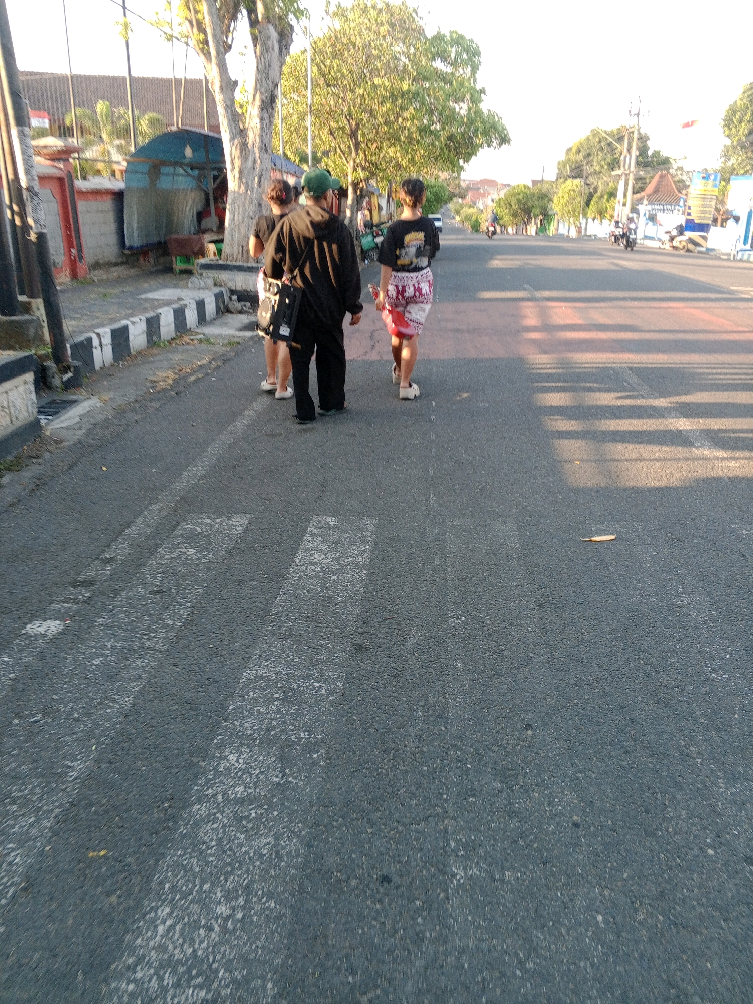The image captures a street scene in Giritirto, Indonesia, with three individuals walking away from the camera. The individuals are on a paved road with a pedestrian crossing marked by white stripes. To the left of the road, there's a sidewalk with a black and white striped curb, a metal pole, and a red and white structure. Further back, a large tree with green foliage shades the area. A blue tarp structure is visible behind the tree. On the right side of the road, trees and buildings can be seen in the distance, along with a few motorbikes and a yellow scooter. The lighting suggests it is late afternoon or early morning, with long shadows cast on the road.