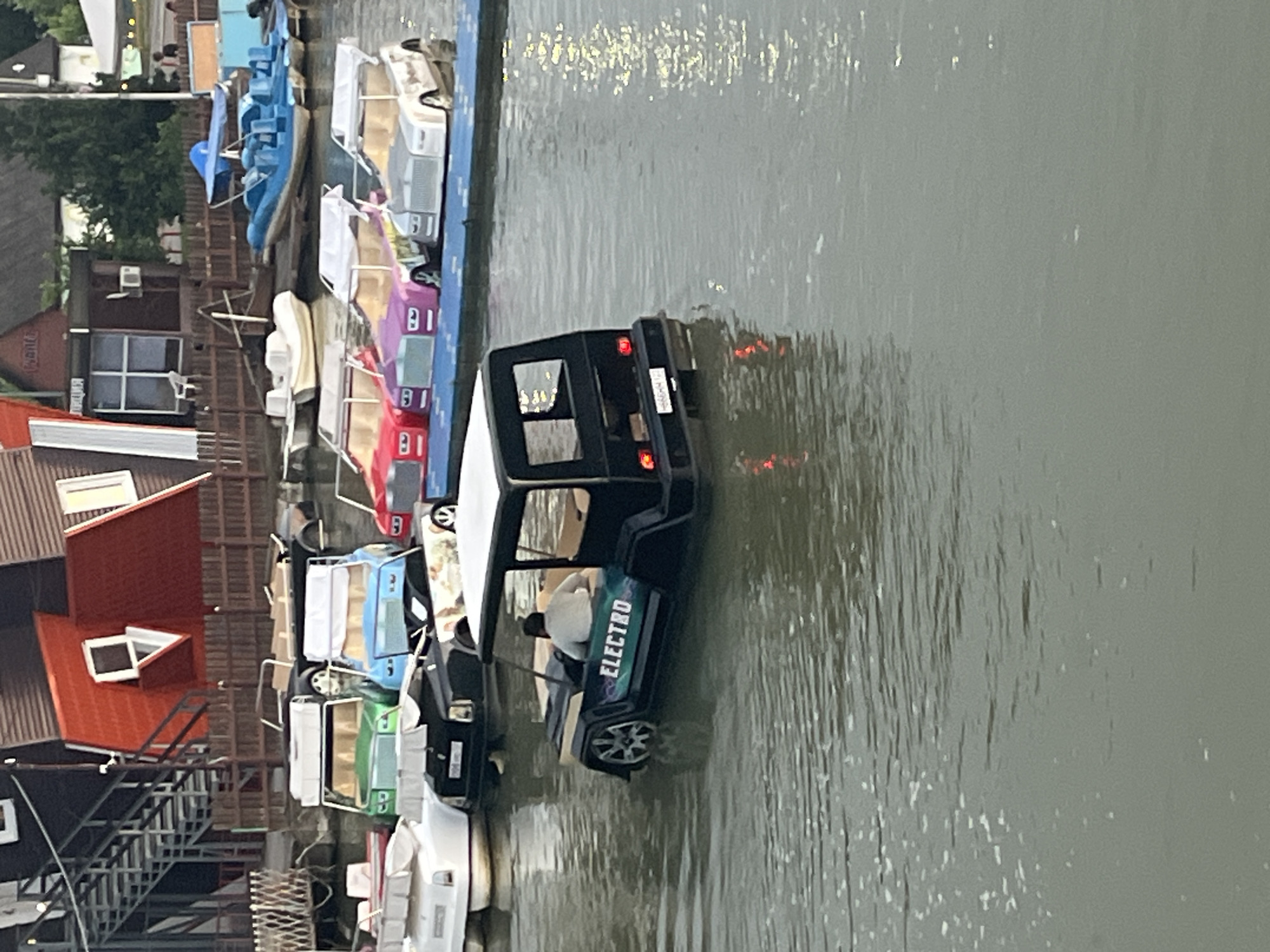The image depicts a body of water, likely a lake or canal, with several amusement park-style boats resembling cars. In the foreground, a black, electric-powered pedal boat with the word "ELECTRO" visible on its side is being operated by a person wearing a light-colored shirt. The boat has a square roof and open sides. In the background, a row of colorful pedal boats, styled after luxury cars in red, purple, and silver, are docked. Further back, several other boats, including a blue inflatable boat and white pedal boats, are visible. The shoreline features wooden fencing, a staircase leading to a red-roofed building, and other structures that suggest a recreational area. The overall scene suggests a warm-weather attraction, possibly a park or resort. This scene is located at the "Europe" shopping and entertainment center in Krasnodar, Russia.