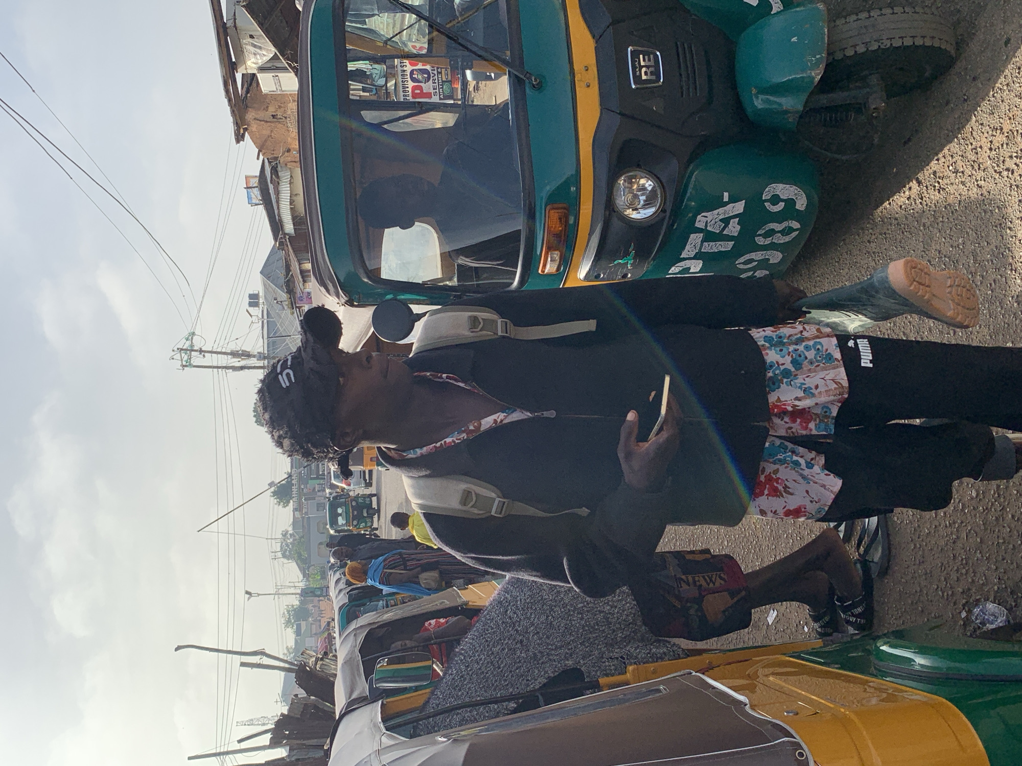 The image captures a street scene in Gura Top, Nigeria, under an overcast sky. In the foreground, a young Black man with dark, curly hair wears a black baseball cap, a black zip-up jacket over a floral-patterned shirt, and black Puma sweatpants. He has a light-colored backpack on and is holding a mobile phone in his right hand. He is carrying a pair of dark green boots in his left hand.
To his right, a green and yellow auto-rickshaw, identified by the license plate "PTA-9989," is parked on the road. The driver, a silhouette of a Black man, is visible through the windshield.
To the left of the foreground subject, another auto-rickshaw, predominantly yellow and white, is partially visible. A Black person, dressed in a gray patterned garment, is seated inside this vehicle. Further in the background, several other auto-rickshaws and cars are on the road. A few other individuals are walking and standing around. Buildings with corrugated metal roofs and wooden structures line the street, indicating a busy urban or semi-urban environment. Overhead, electrical wires crisscross the sky. The ground is paved with asphalt.