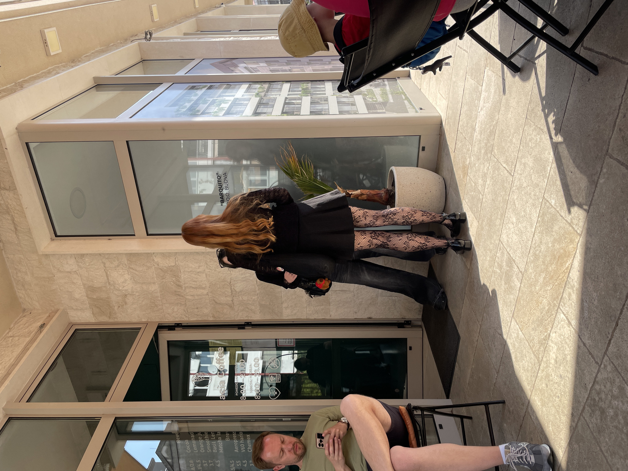 The image depicts an outdoor scene in Budva, Montenegro, outside what appears to be a cafe or shop on a sunny day. In the mid-ground, a couple is embracing near a glass storefront. The woman has long, reddish-brown hair, a black dress, black patterned tights, and black boots. The man, wearing dark jeans and a black top, holds a small plush toy in his hand.
To the left, a bearded man in an olive green t-shirt, shorts, and grey sneakers is seated on a black outdoor chair, looking at his mobile phone. His legs are extended, resting casually. Partially visible in the upper right, another person wearing a yellow hat and a red top is seated at a black table.
The setting includes a light-colored stone tiled pavement, a building with a textured stone facade, and large glass windows reflecting the surroundings. A large potted plant with palm-like leaves stands next to the embracing couple. Strong shadows are cast across the pavement, suggesting bright daylight.
Visible text on the glass storefront includes the name "Acia Coffee" and operating hours: "PONEDELJAK-PETAK 8:00-21:00" (Monday-Friday) and "SUBOTA-NEDJELJA 8:00-21:00" (Saturday-Sunday). A menu is also displayed, listing beverages such as Cocoa, Lemonade, Orange juice, Smoothie, Basil Lemon, Matcha latte, Brew, Aeropress, V60 hario, and Chemex, along with their prices. Another sign clearly reads "BARQUITO" O.O. BUDVA.