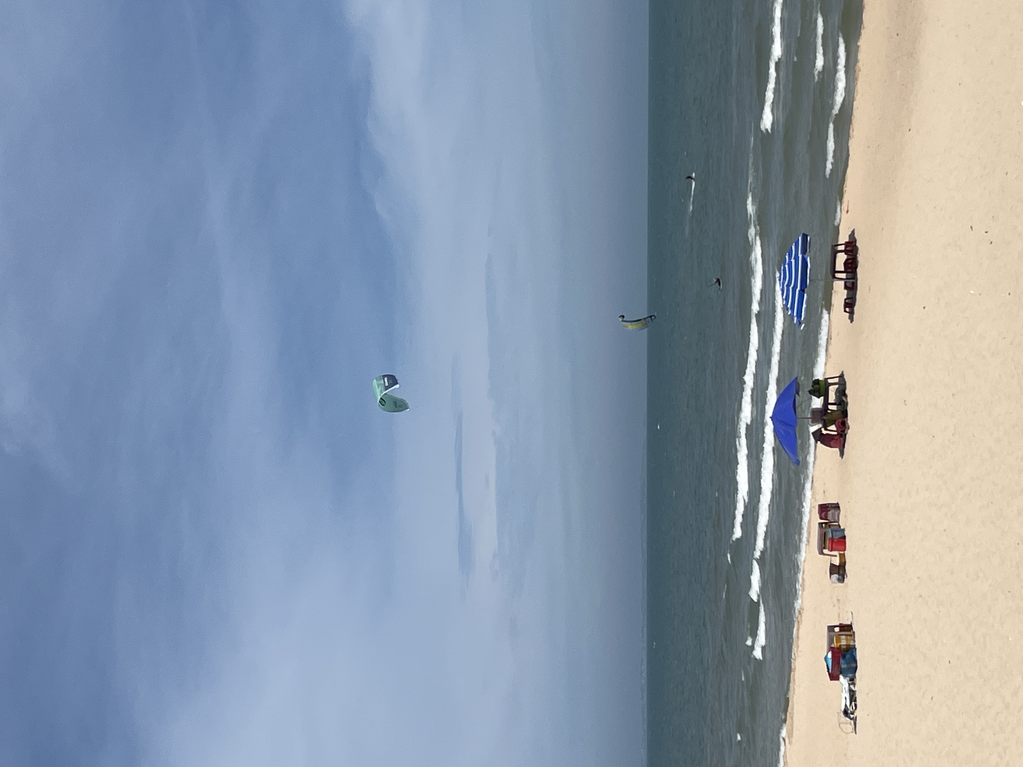The image depicts a wide expanse of a sandy beach under a partly cloudy blue sky. The sea is a deep blue-green, with moderate waves breaking near the shore. In the distance, there are faint outlines of land visible, likely the coastline. The sky is dominated by pale blue with scattered white clouds.
Several kitesurfers are present in the water. One large, light green kite is high in the sky towards the left of the frame, with a kitesurfer attached. Another, smaller kite, yellow and blue, is lower in the sky, also with a kitesurfer. Further out to sea, another kitesurfer can be seen on the water.
On the beach, two umbrellas are set up. The one on the left is a solid blue umbrella, with a table and chairs underneath, where two figures appear to be seated. On the right, a blue and white striped umbrella covers a table with several chairs. In front of these setups, there are various containers and bags on the sand. Closer to the left, a few colorful plastic bins are stacked together.
The beach itself is a light beige sand, appearing smooth and somewhat wet near the water's edge. The overall scene suggests a recreational day at the beach, with activities like kitesurfing and relaxation under umbrellas. The location is identified as Phu Thuy Ward, Vietnam.