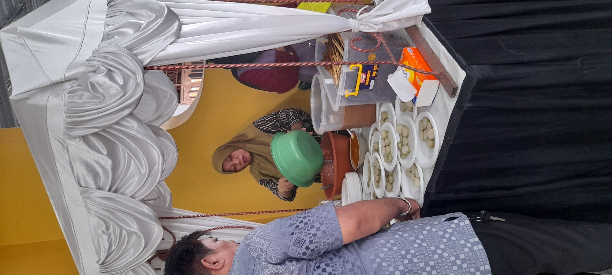 The image depicts a food stall set up under a white draped tent, likely at an outdoor event in Taman, Indonesia. A woman wearing a brown hijab and a patterned shirt is pouring something from a large green bowl into a reddish-brown basin. In front of her, a table is laden with white paper plates, each holding several pieces of what appear to be food items, possibly snacks or appetizers. To the right, a person in a dark-colored top and a purple vest is partially visible, also near the food display. On the table, there are also large plastic containers, one filled with skewers and another with a brown liquid. A box of tissues is also visible. A man in a blue patterned shirt is standing to the left, facing the table and seemingly observing the food. The background is a bright yellow wall, and the stall itself has a black draped skirt around the table. The overall scene suggests a food service operation at a local gathering or market.